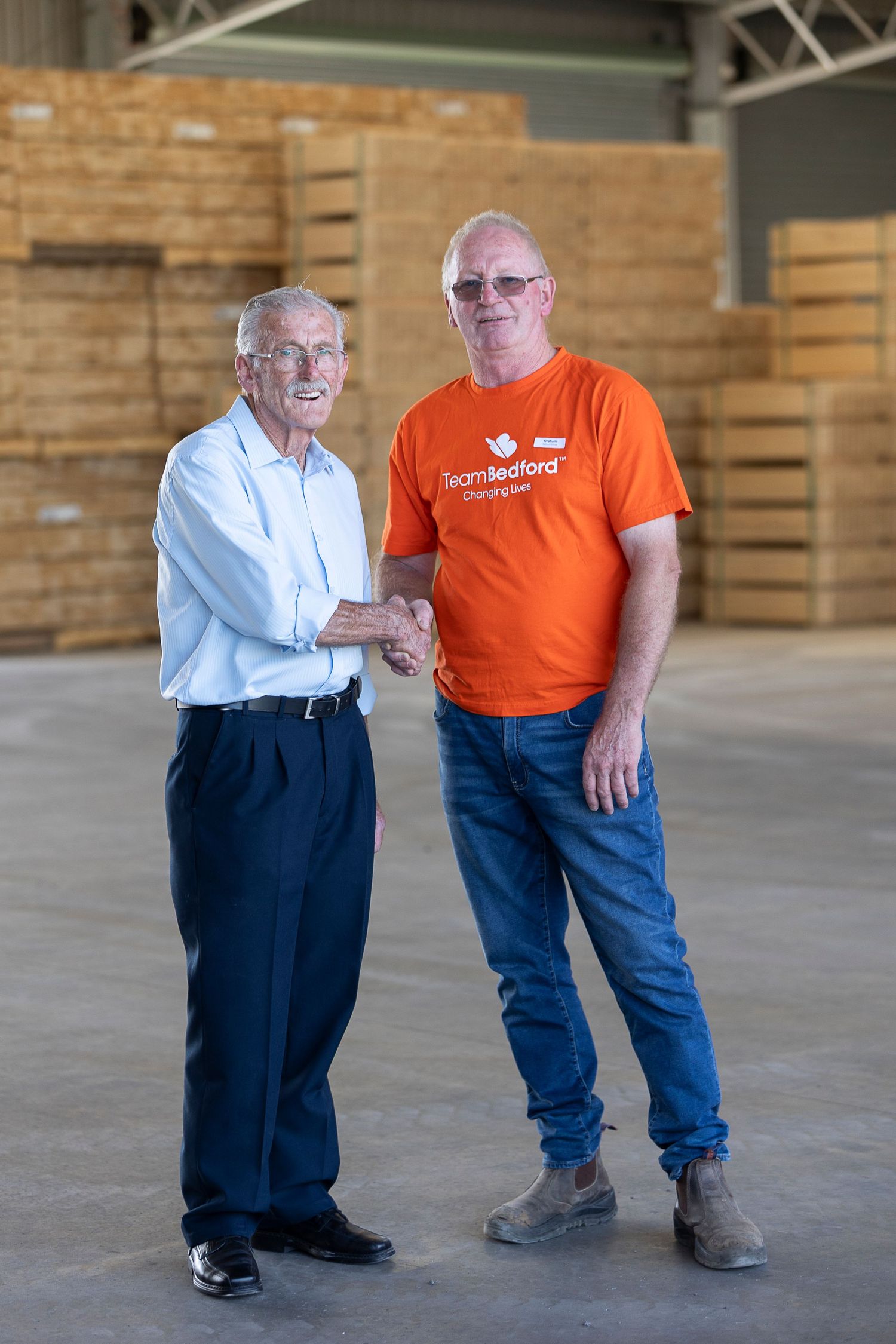 Two people stand in a warehouse, one wearing a white shirt and the other in an orange t-shirt.