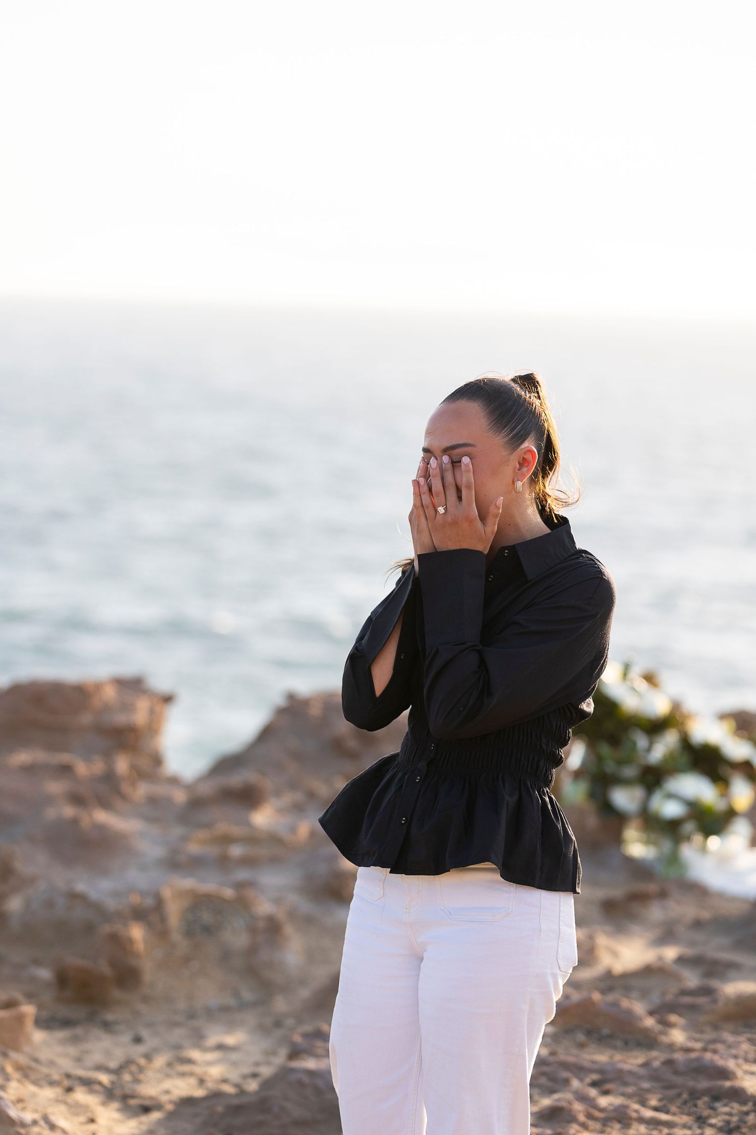 An emotional surprise proposal moment captured at sunset on a rocky oceanfront cliff.