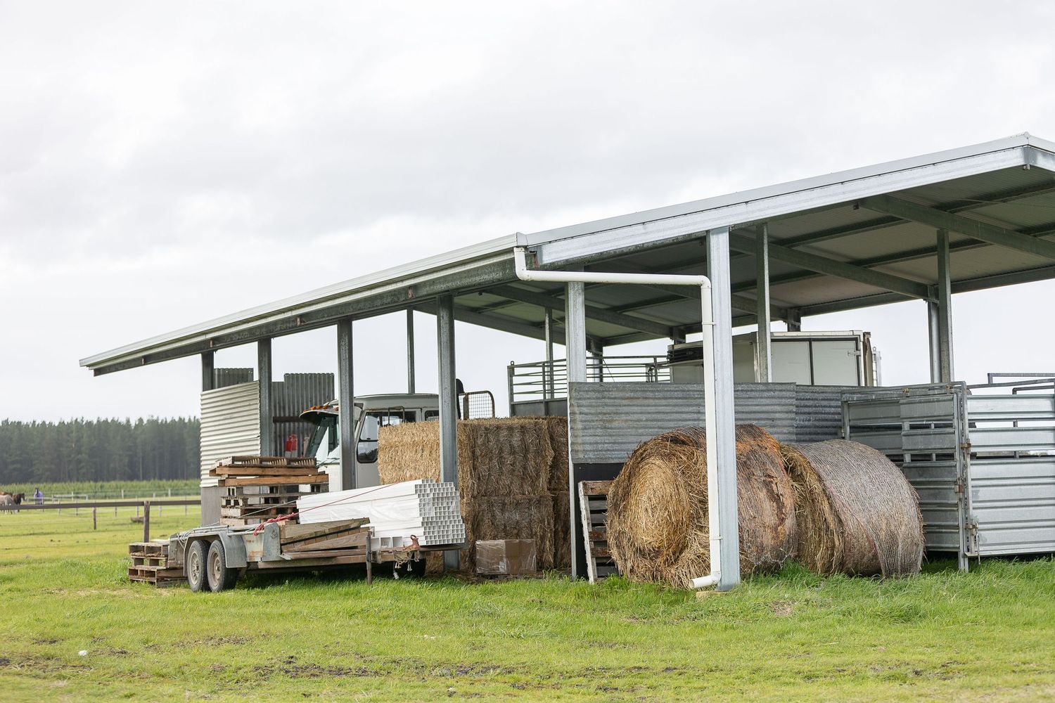 Farm shed with hay bales, equipment, and trailer on green pasture under overcast sky.