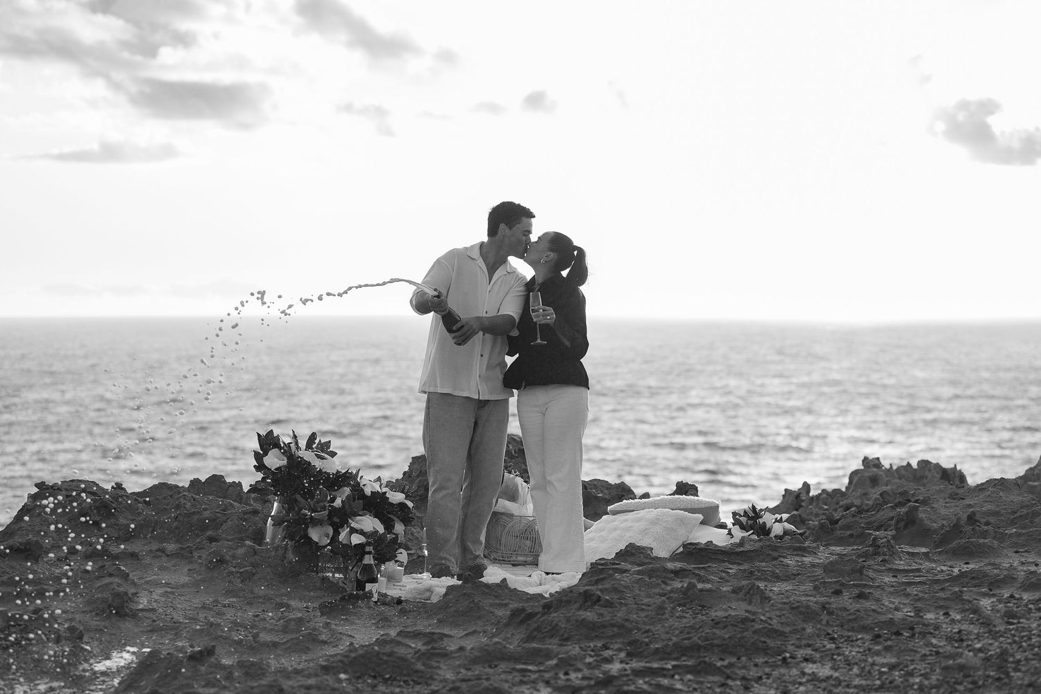 Silhouetted couple sitting on rocky coastline at sunset.