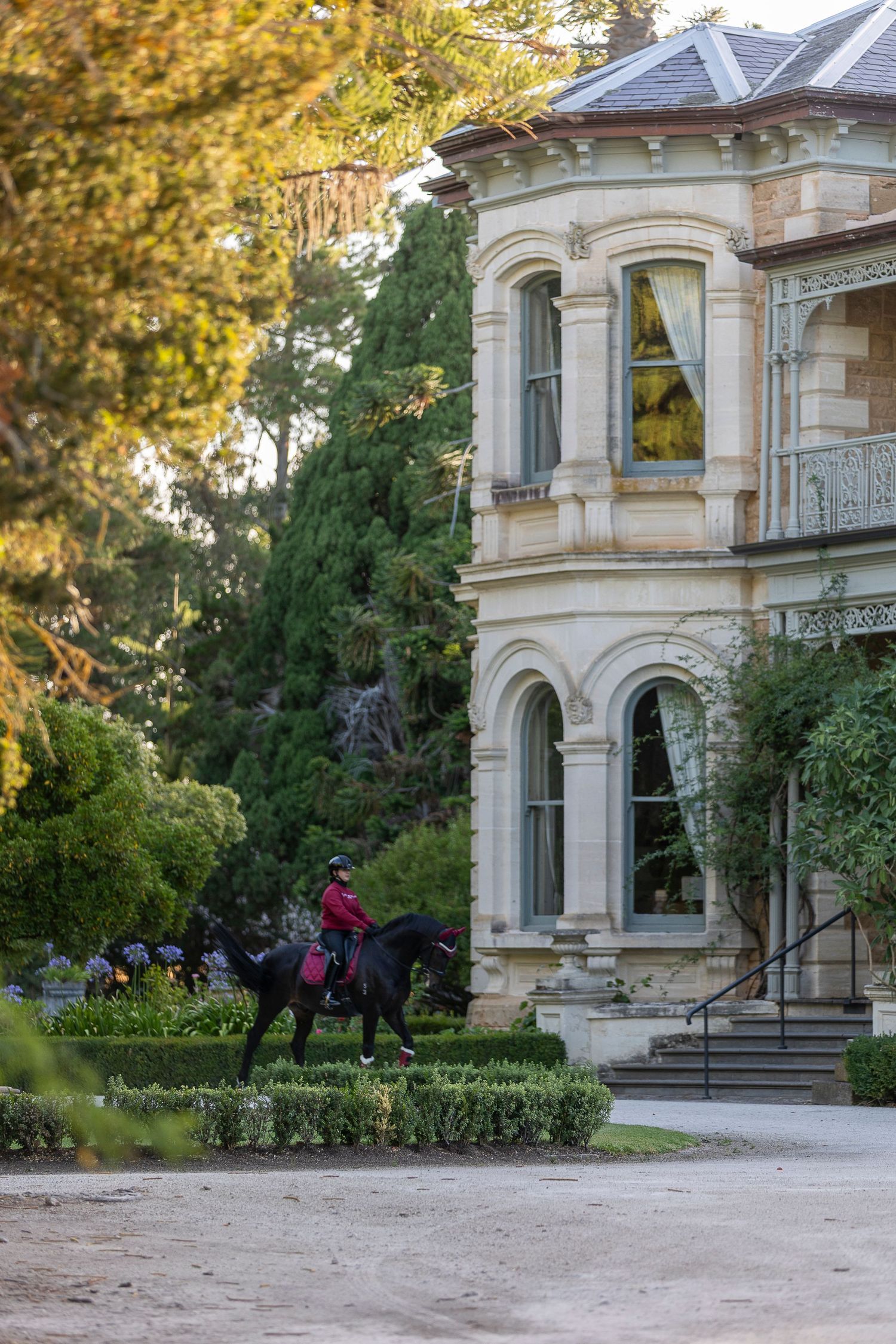 Person riding a dark horse beside a grand Victorian mansion surrounded by lush gardens and tall trees.