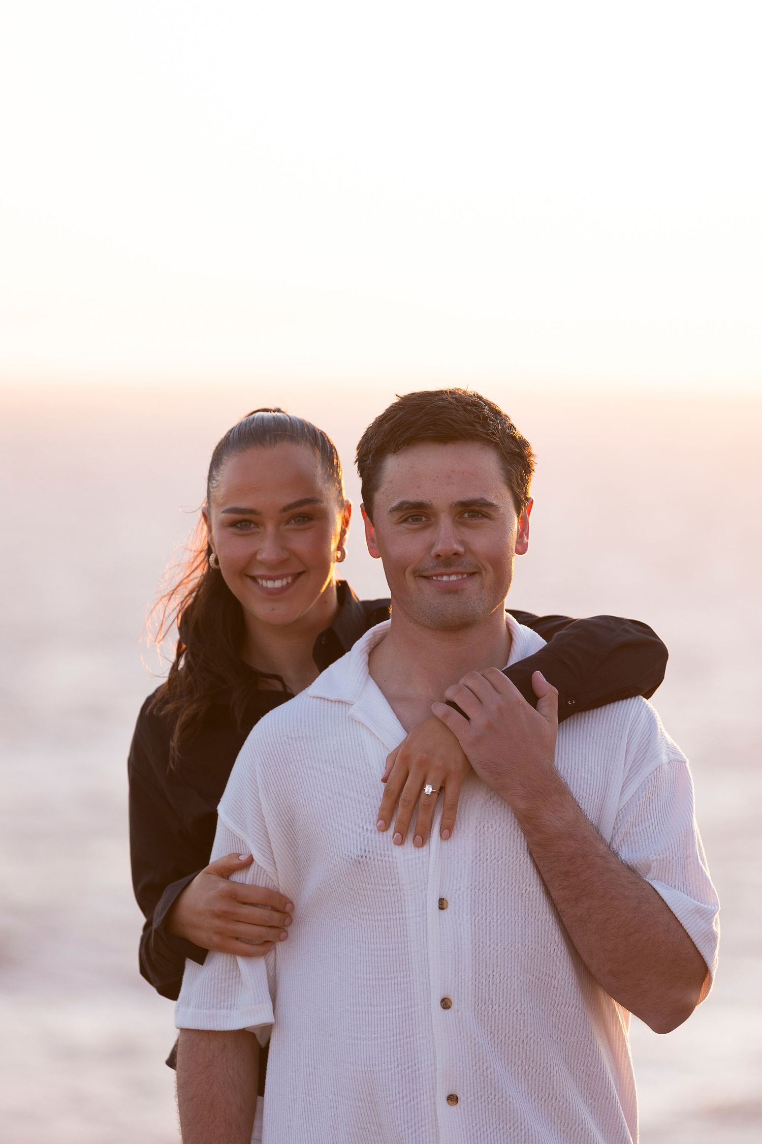 A couple embraces and smiles while posing on a beach at sunset.