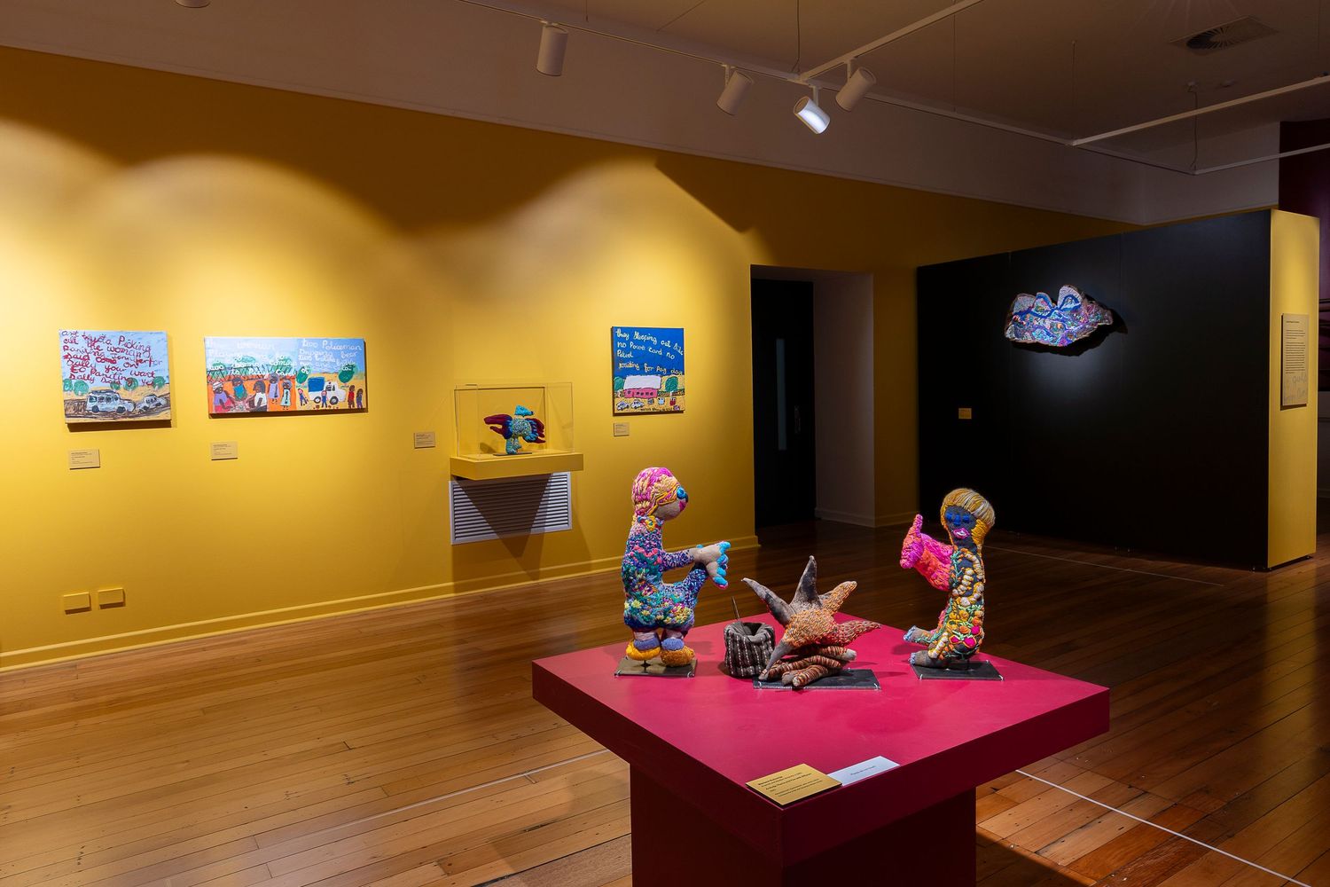 Art gallery space with yellow walls displaying framed photographs and sculptures on red pedestals.