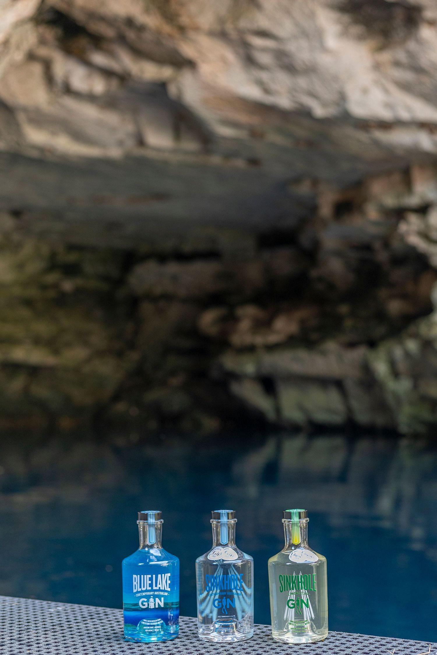 Three gin bottles reflected in still blue water against a rocky cave backdrop.