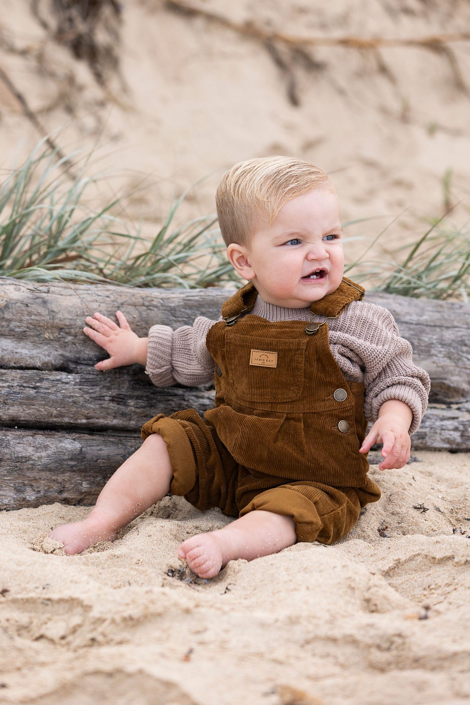 A baby in brown corduroy overalls sits playfully on driftwood at the beach.