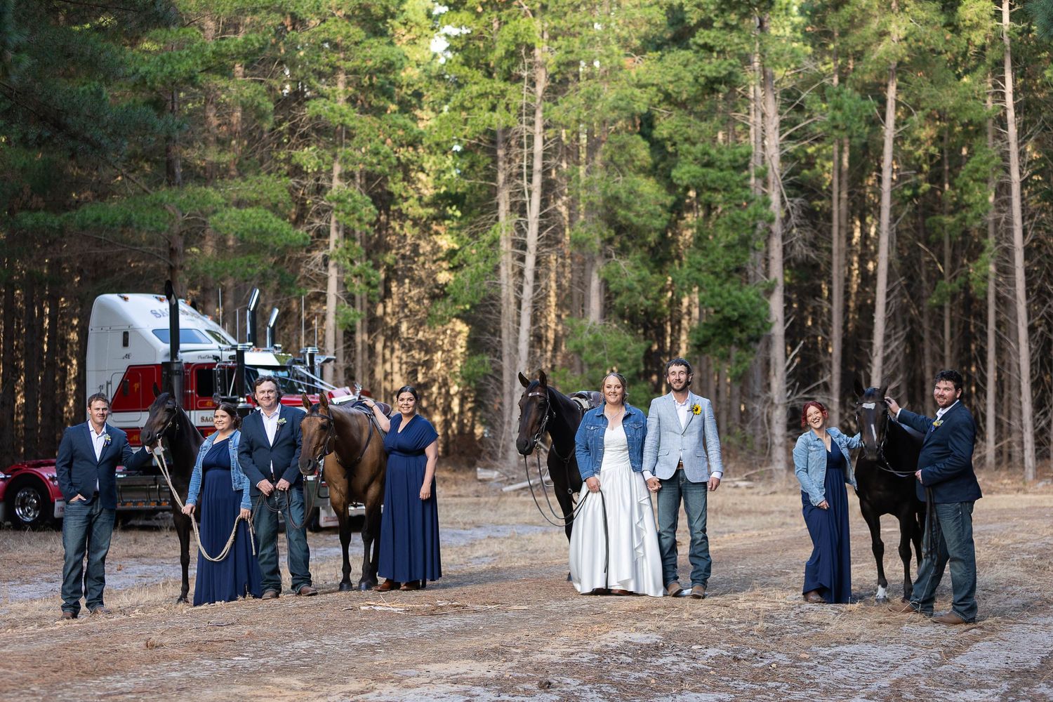 Wedding party gathered in a pine forest clearing wearing navy blue and white formal attire.