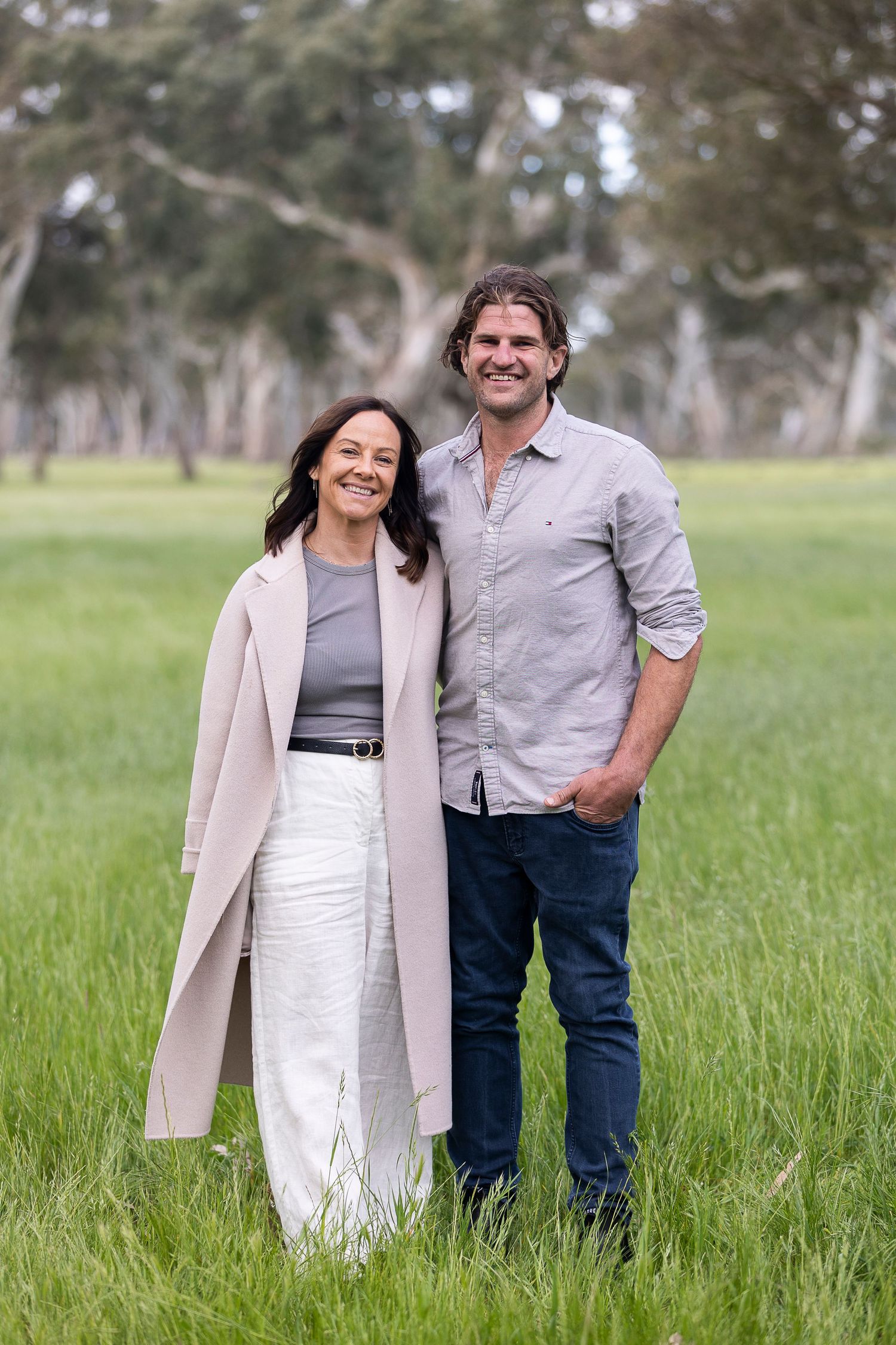 Couple in casual attire poses together in a grassy field on an overcast day.