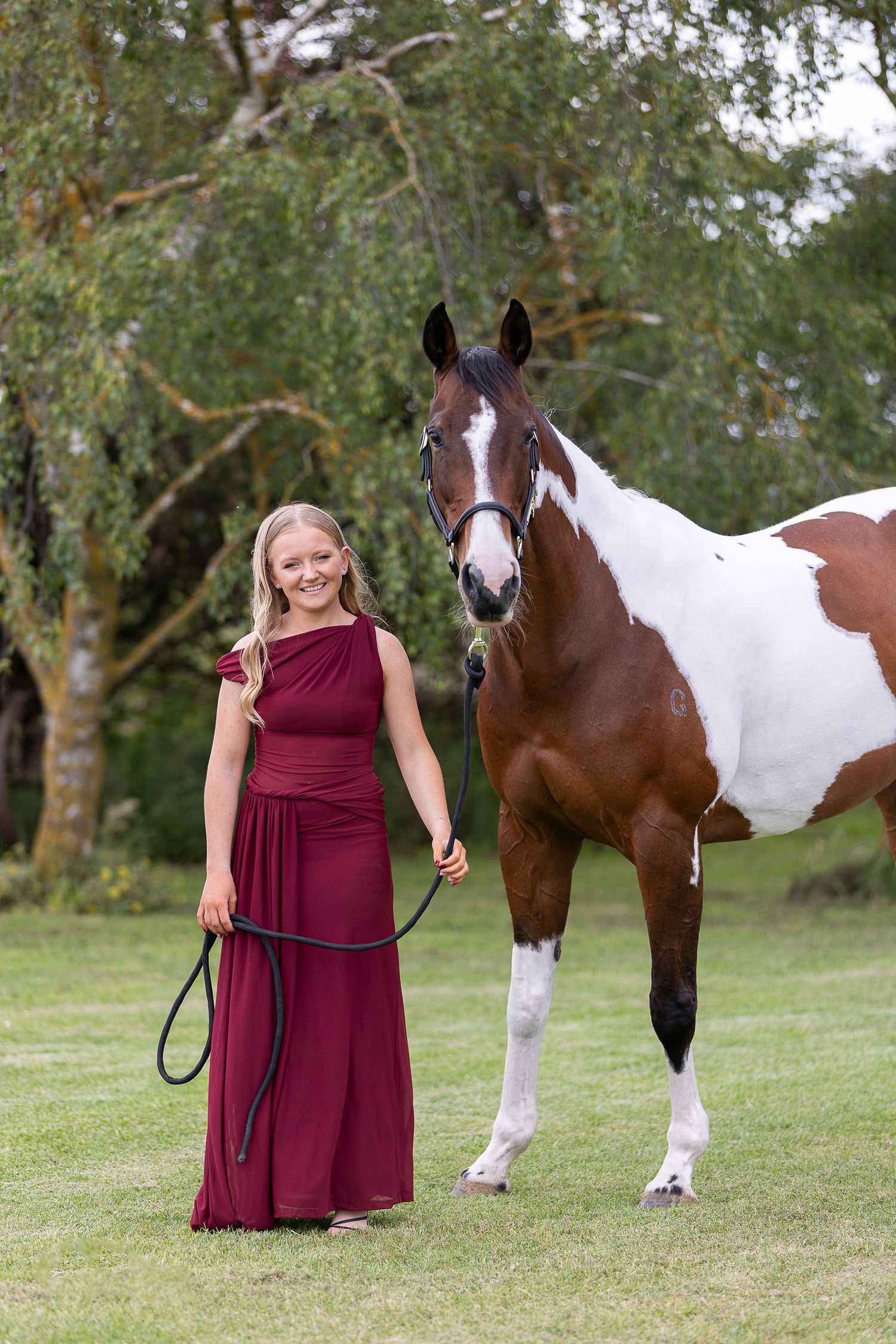A paint horse and person in burgundy dress stand together in a grassy field.