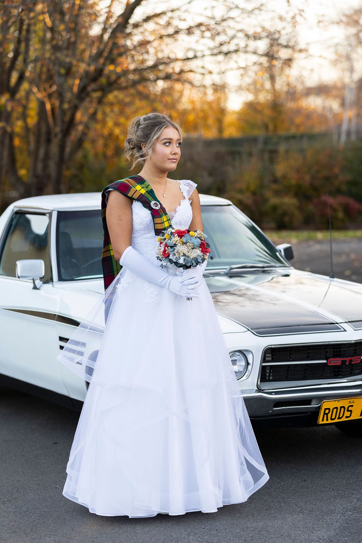 A bride in a white gown poses beside a classic white GTO muscle car during autumn.
