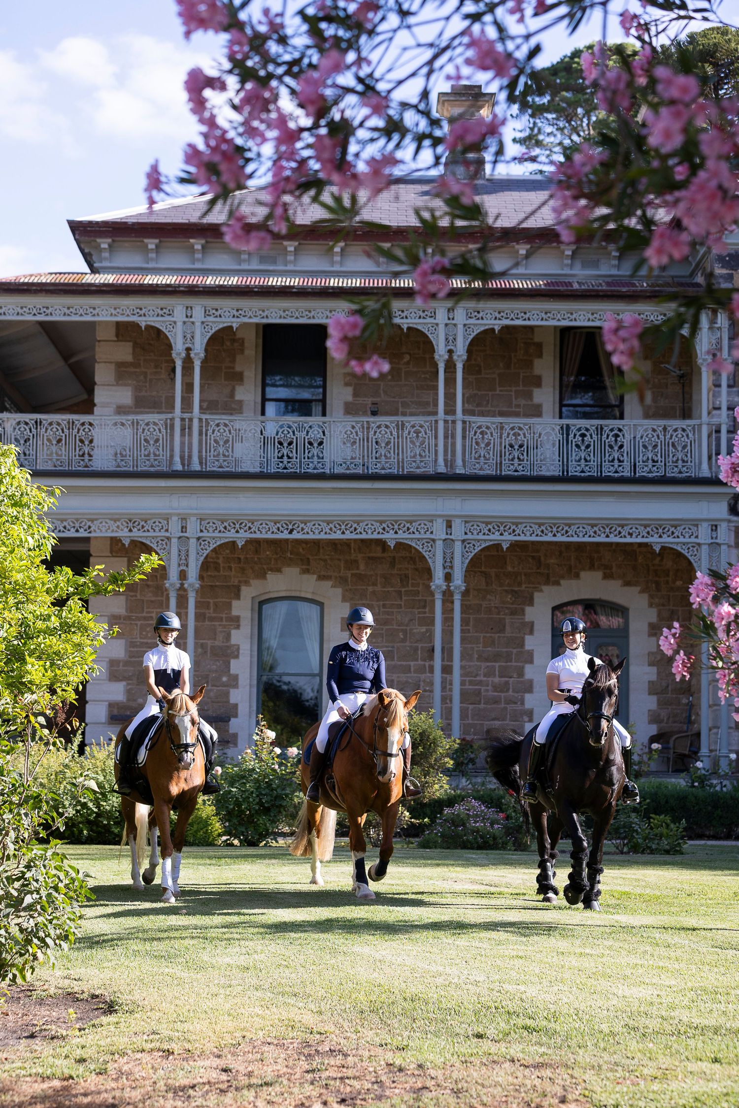 Horses and riders traverse across a manicured lawn in front of a historic stone building with ornate trim.