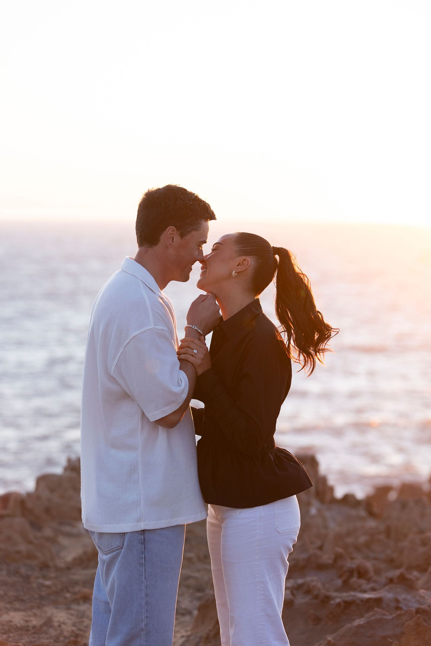 A couple in contrasting outfits embraces on a rocky beach at sunset.