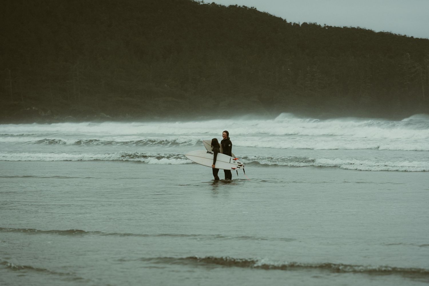 Silhouettes of beachgoers stand in the distance against crashing waves and an overcast sky