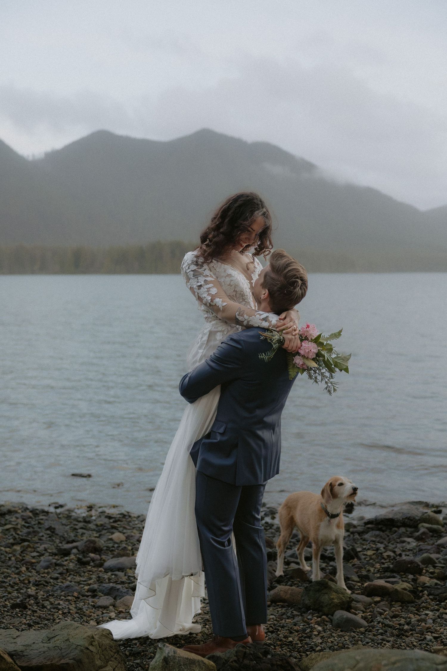 A couple embraces on a rocky beach with their dog watching nearby and mountains in the background.
