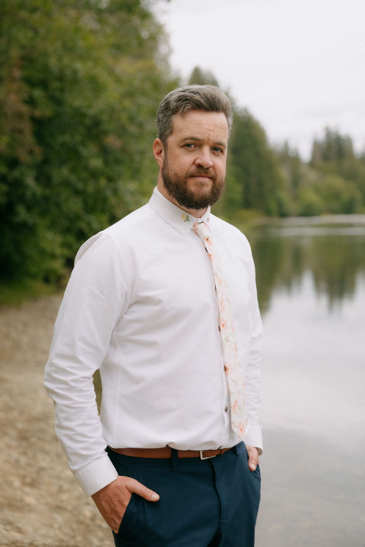 Bearded man in white shirt and floral tie standing by a serene lake surrounded by lush green trees.