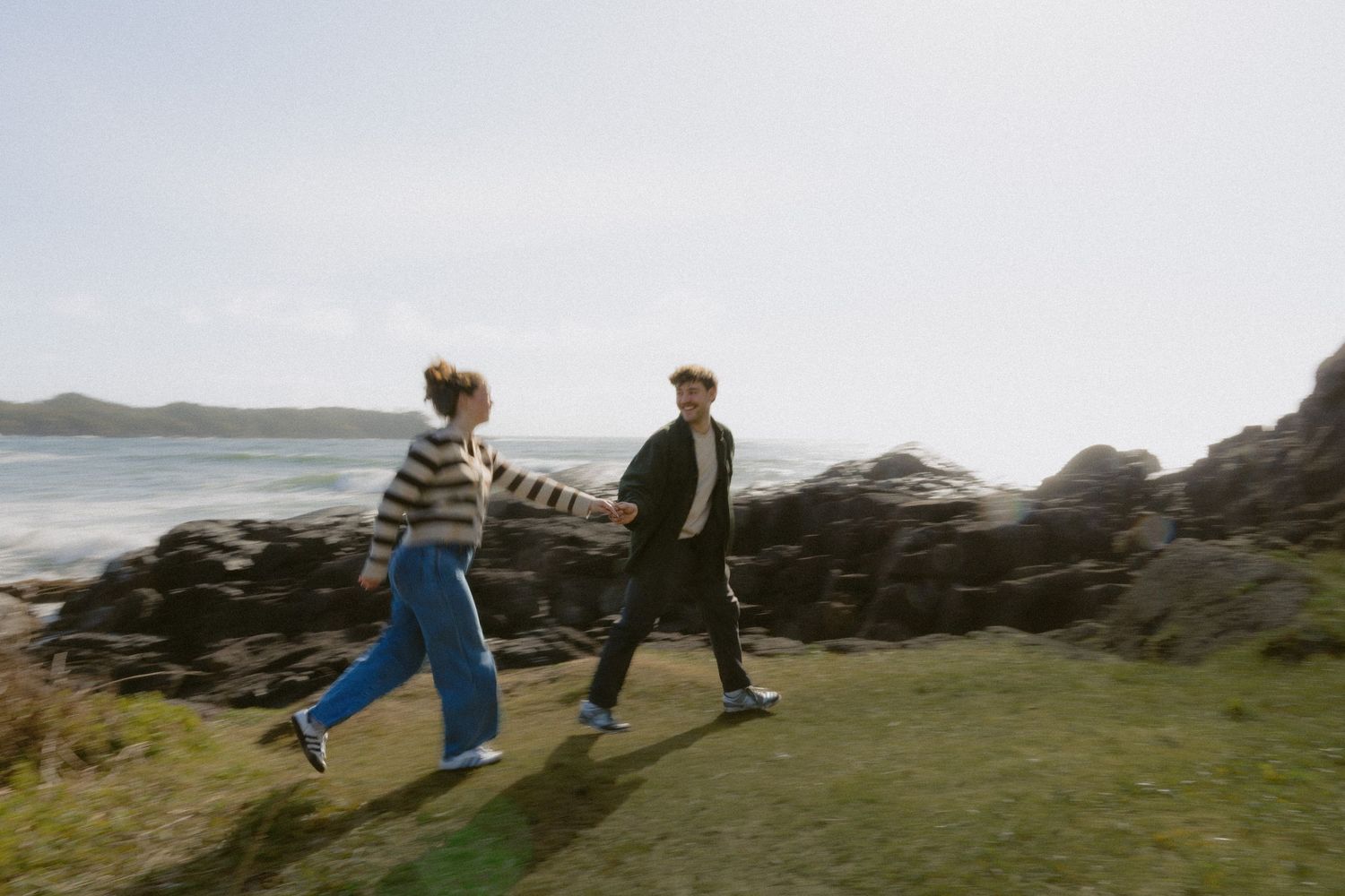 A couple walks hand in hand along a scenic coastal path with rocky shores and ocean views.