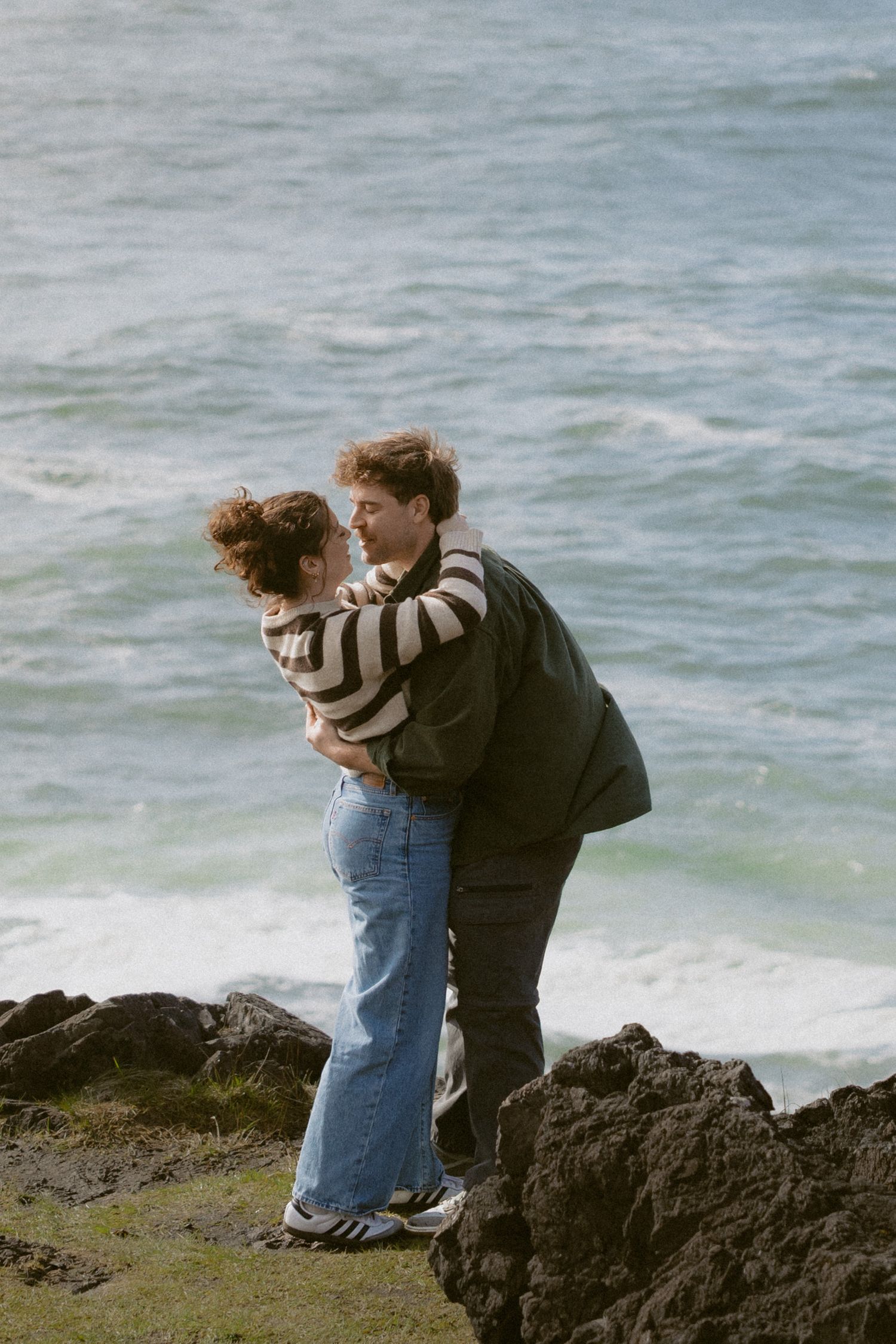 Romantic couple embracing and almost kissing on rocky seaside cliffs with turquoise ocean waves behind them.