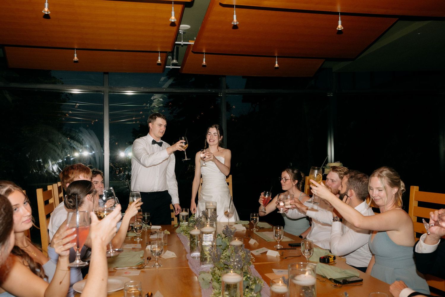 Bride and groom toast with guests at elegant indoor wedding reception dinner table with candles.