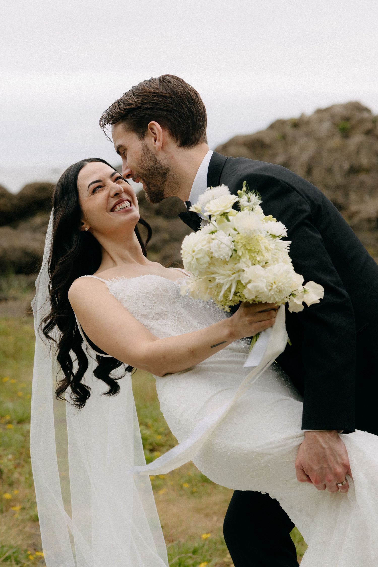 Groom kisses bride's cheek as she laughs holding white floral bouquet in outdoor wedding portrait.