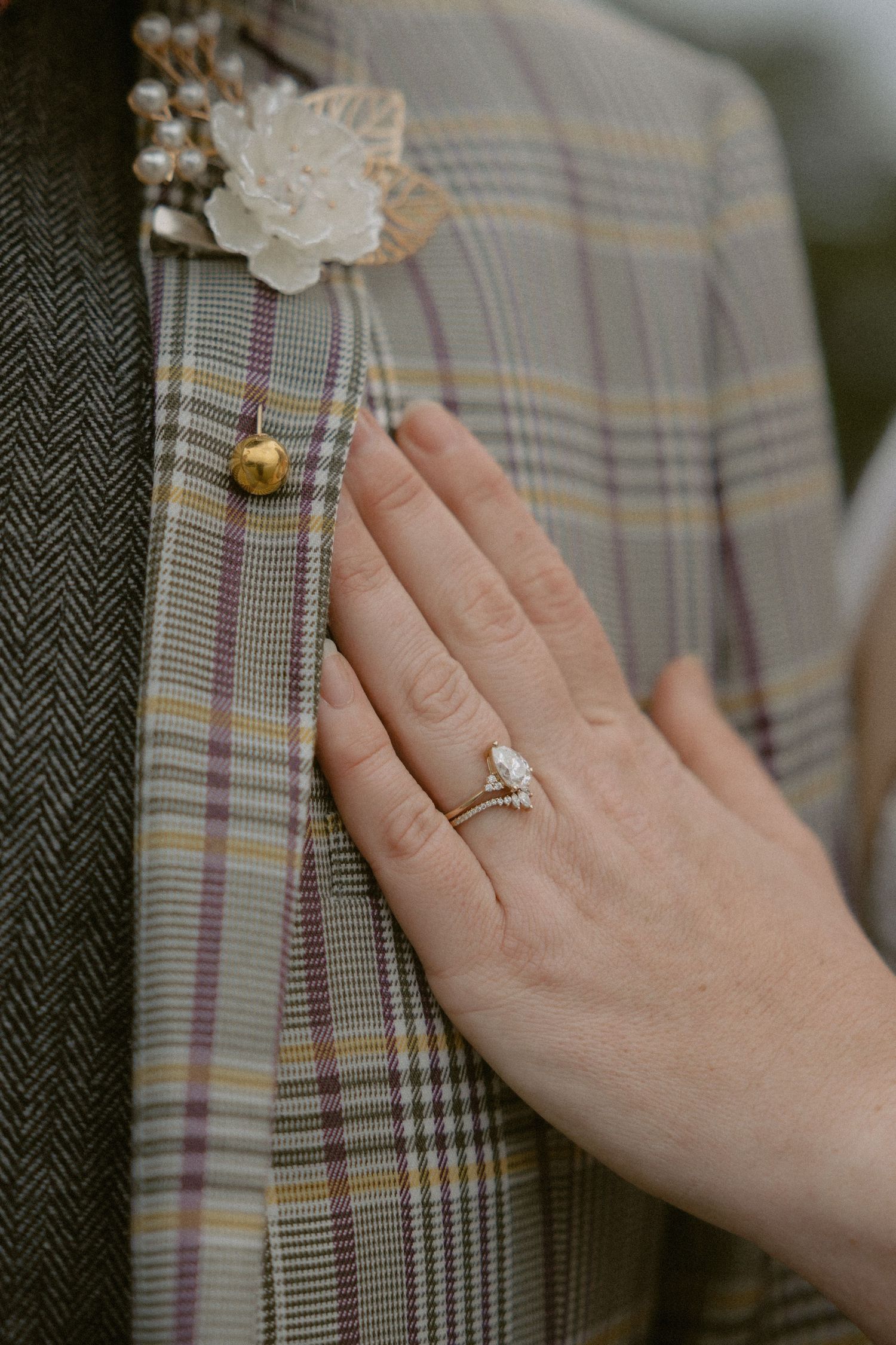 Close up of an engagement ring and plaid suit jacket with floral boutonniere.