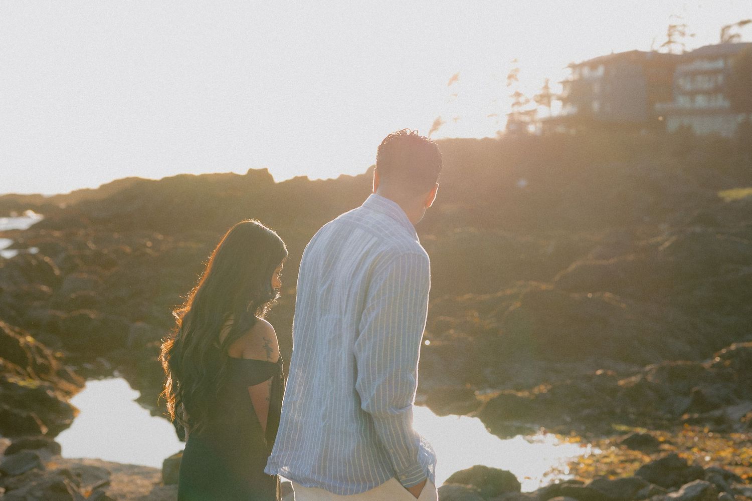Couple standing together at golden hour near rocky coastline with warm sunset light glowing behind them.