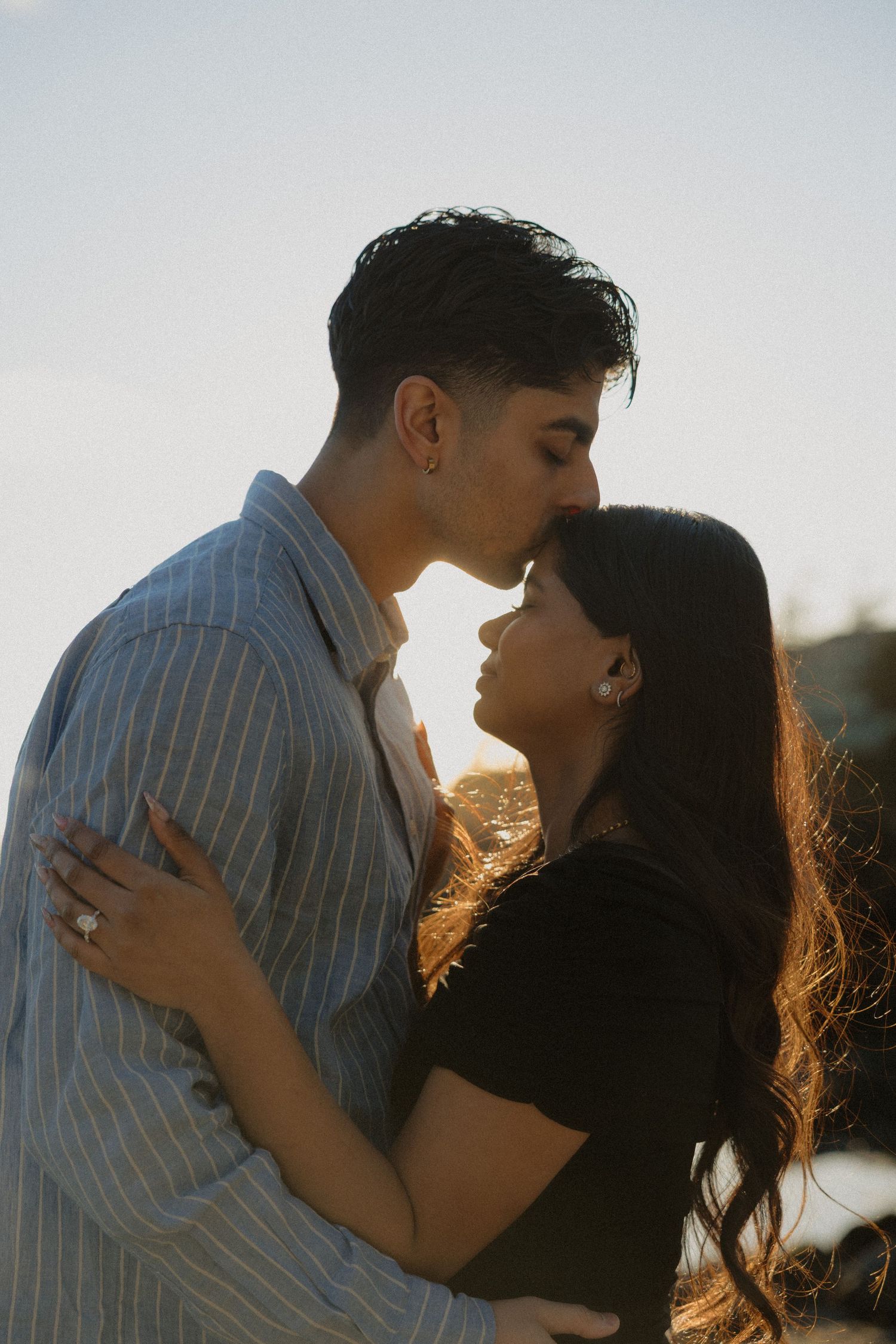 Romantic couple sharing a kiss at sunset, backlit by warm golden light on a rocky coastal beach.