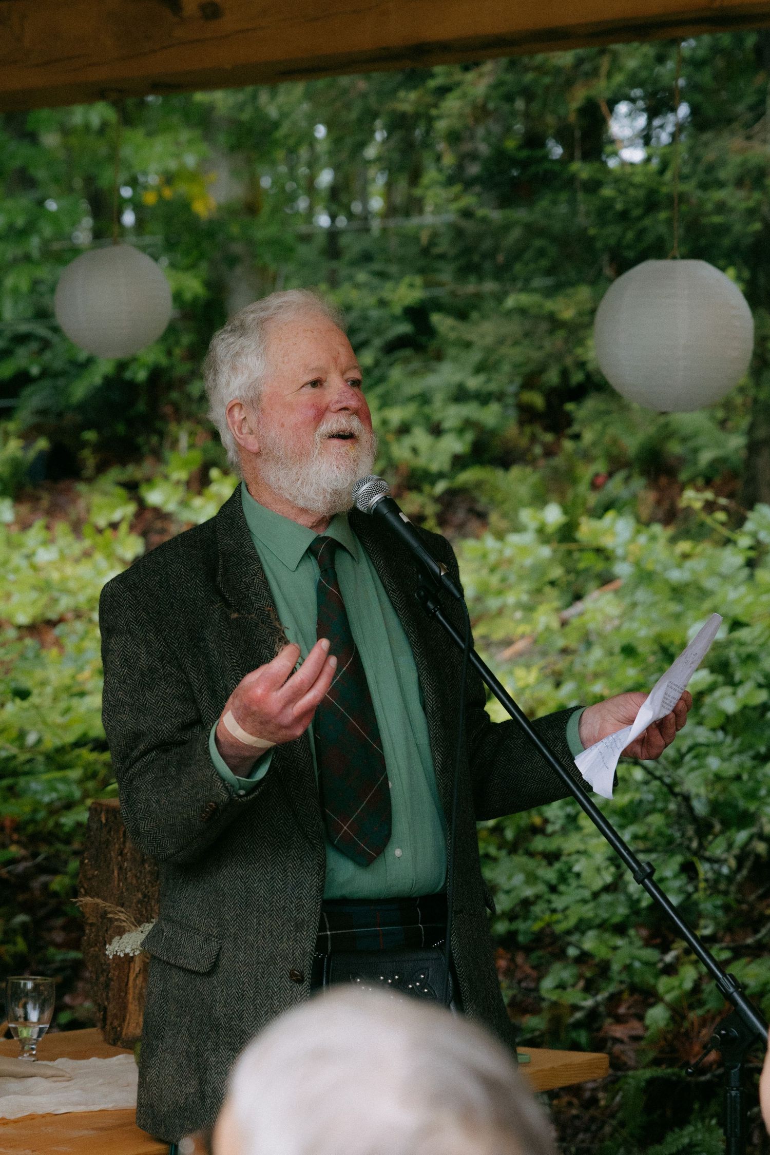 Elderly bearded man in green shirt and plaid tie speaking at outdoor microphone during garden event.