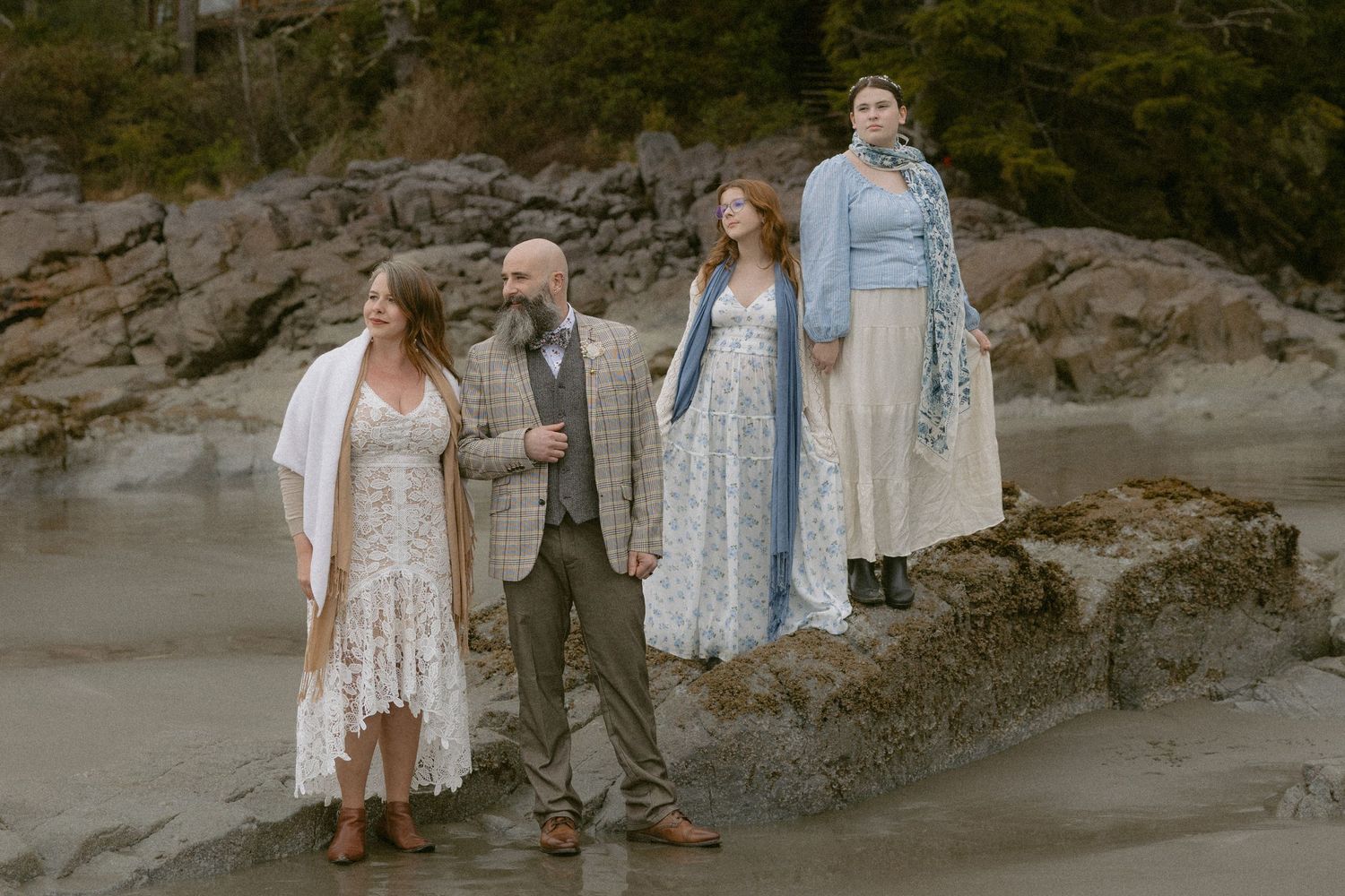 A group stands on rocky coastal terrain wearing light colored flowy dresses and formal attire.