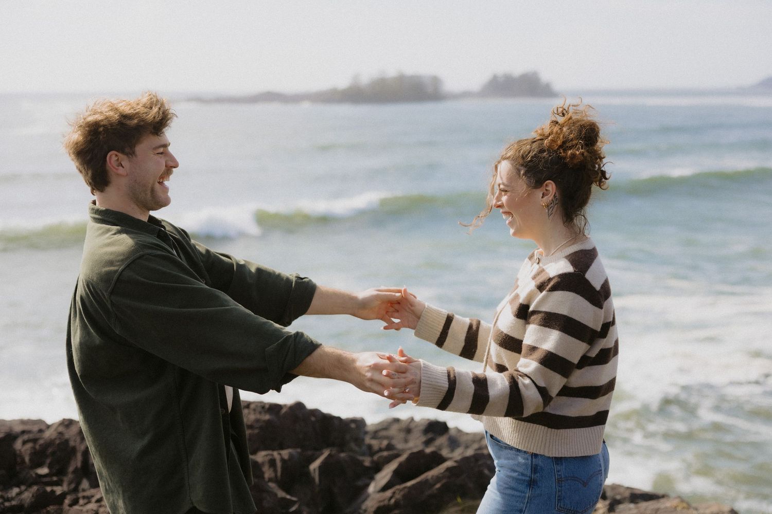 Happy couple holding hands and dancing on a rocky beach with ocean waves in the background.