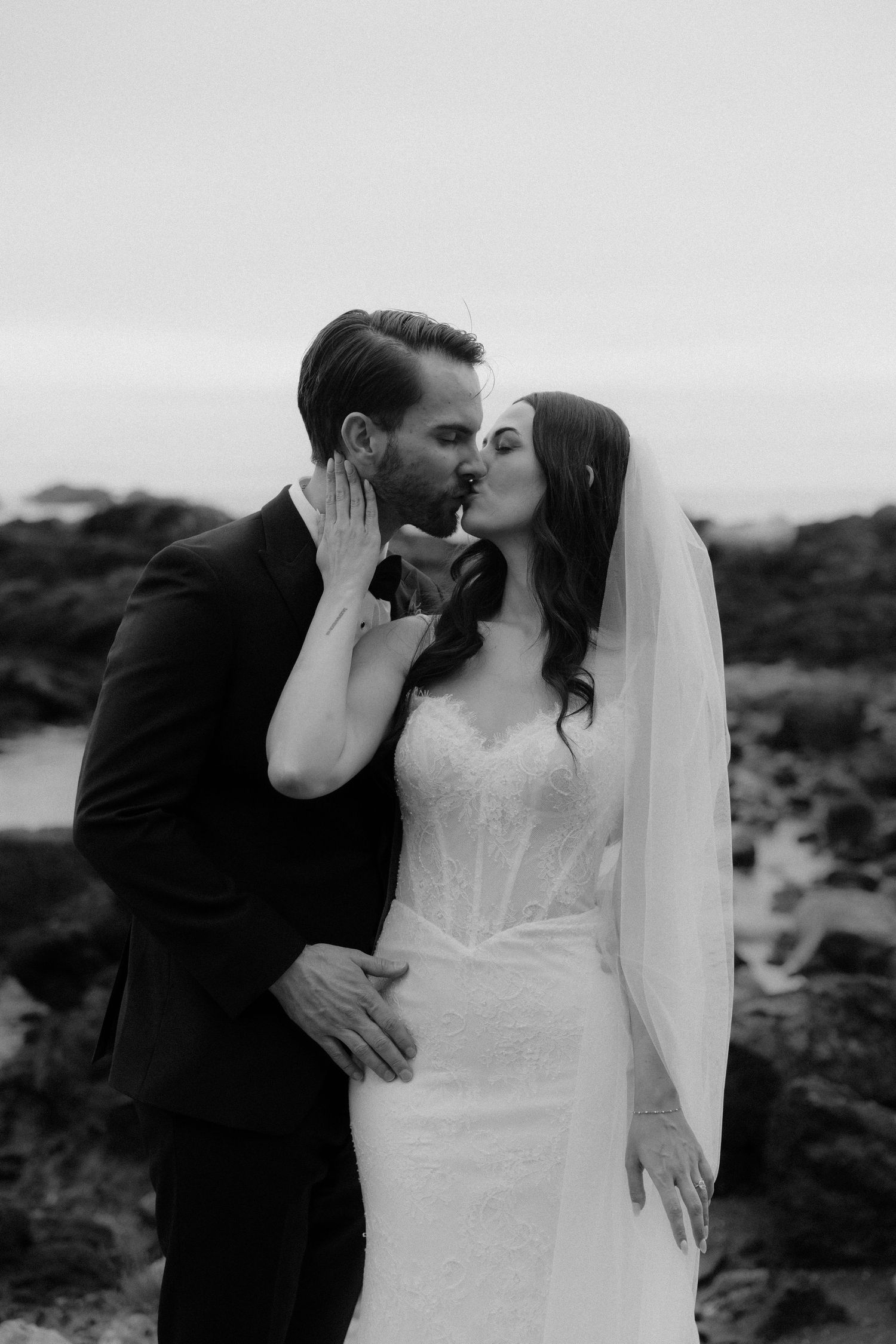 Black and white photo of a bride and groom sharing a romantic kiss outdoors, bride in lace gown with veil.
