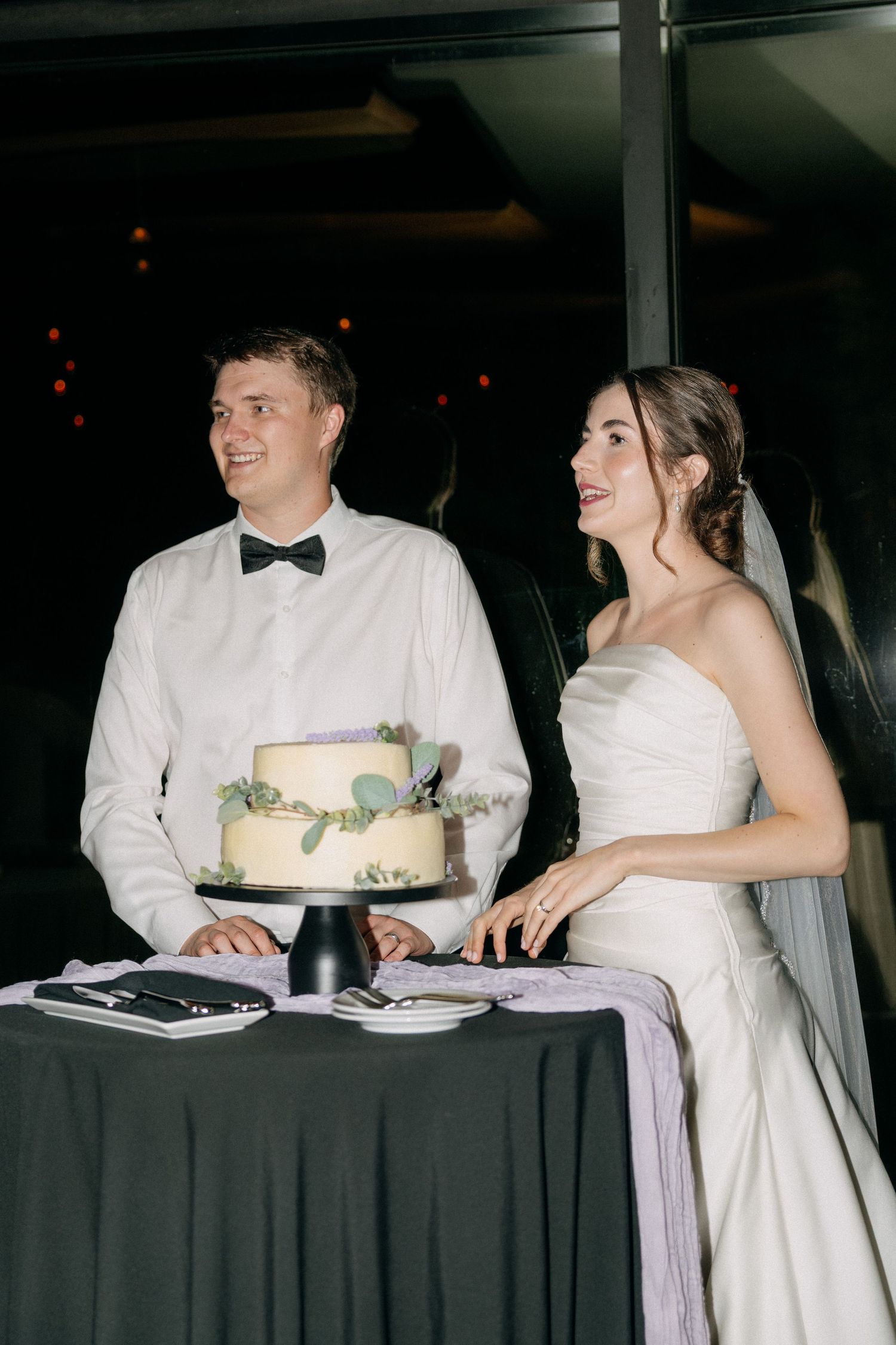 Smiling bride and groom standing together at a table with a two-tier white wedding cake at their reception.