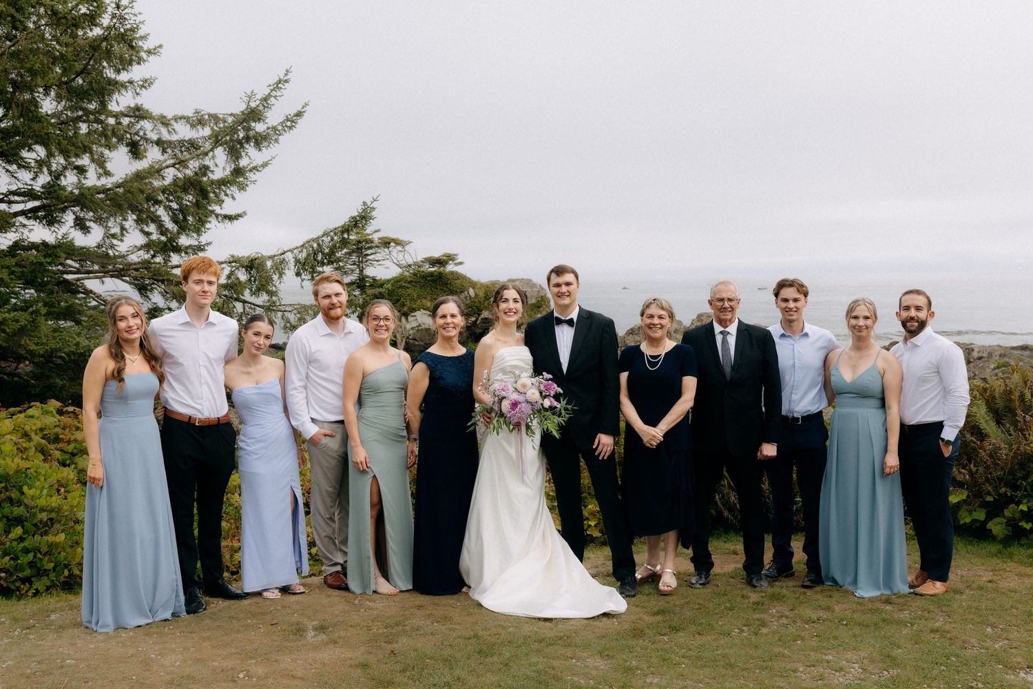 Wedding party posing outdoors with bride and groom, bridesmaids in blue dresses, ocean view backdrop.