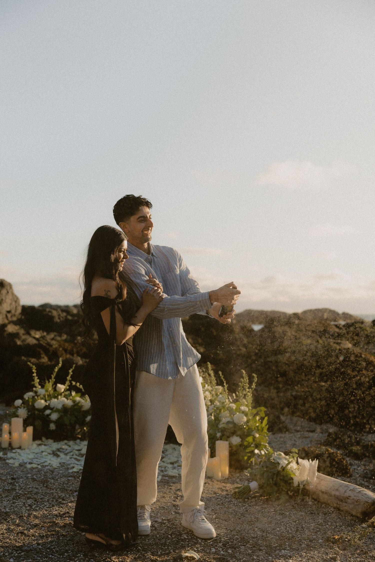 Couple dancing at sunset on rocky beach with candles and floral arrangement during romantic proposal scene.