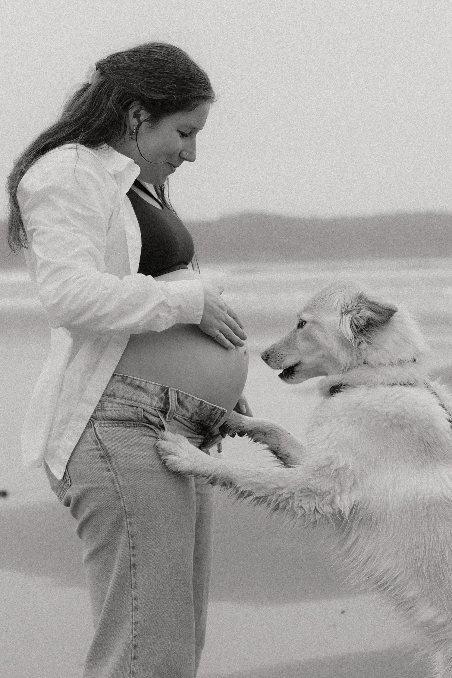 A pregnant woman in a white shirt and jeans interacts lovingly with her white dog on a beach in black and white.