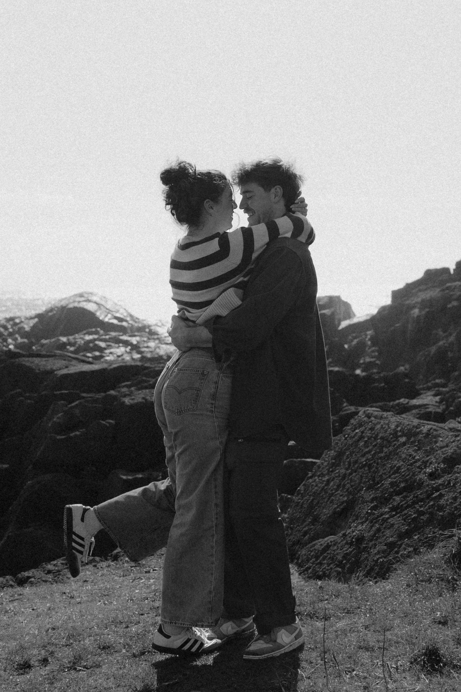 Black and white photo of a couple embracing romantically on rocky coastal cliffs with misty mountains behind.