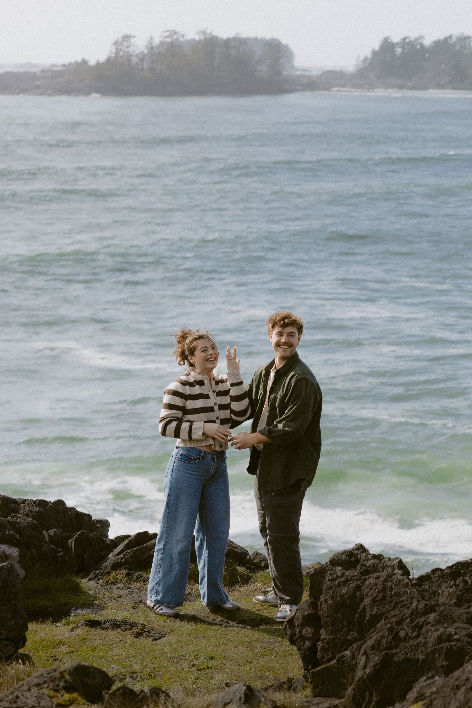 Couple smiling and waving on rocky coastal cliffs with turquoise ocean waves in the background.