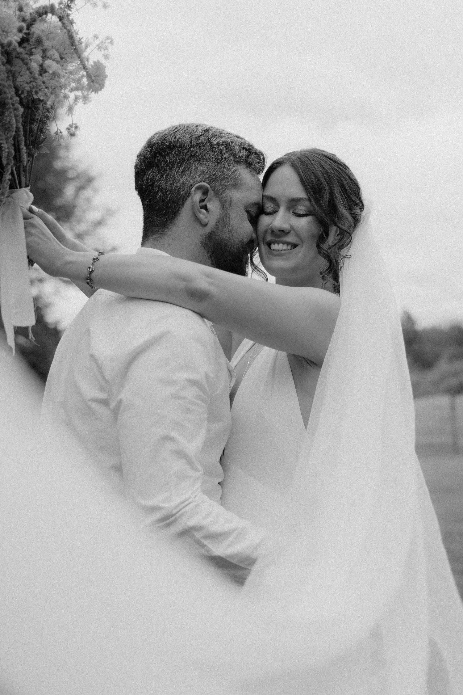 Black and white photo of a smiling bride and groom sharing a tender moment, with veil flowing and bridal bouquet overhead.