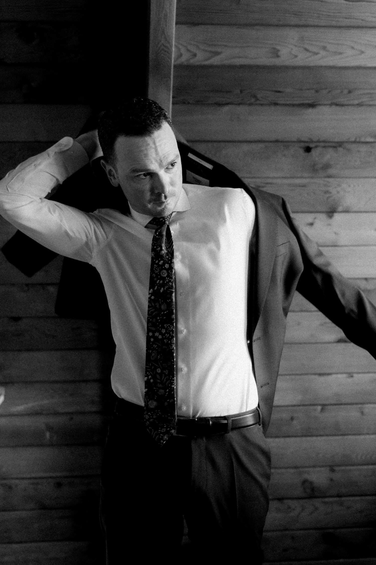Black and white photo of a man in a suit and tie putting on his jacket against a wooden wall background.