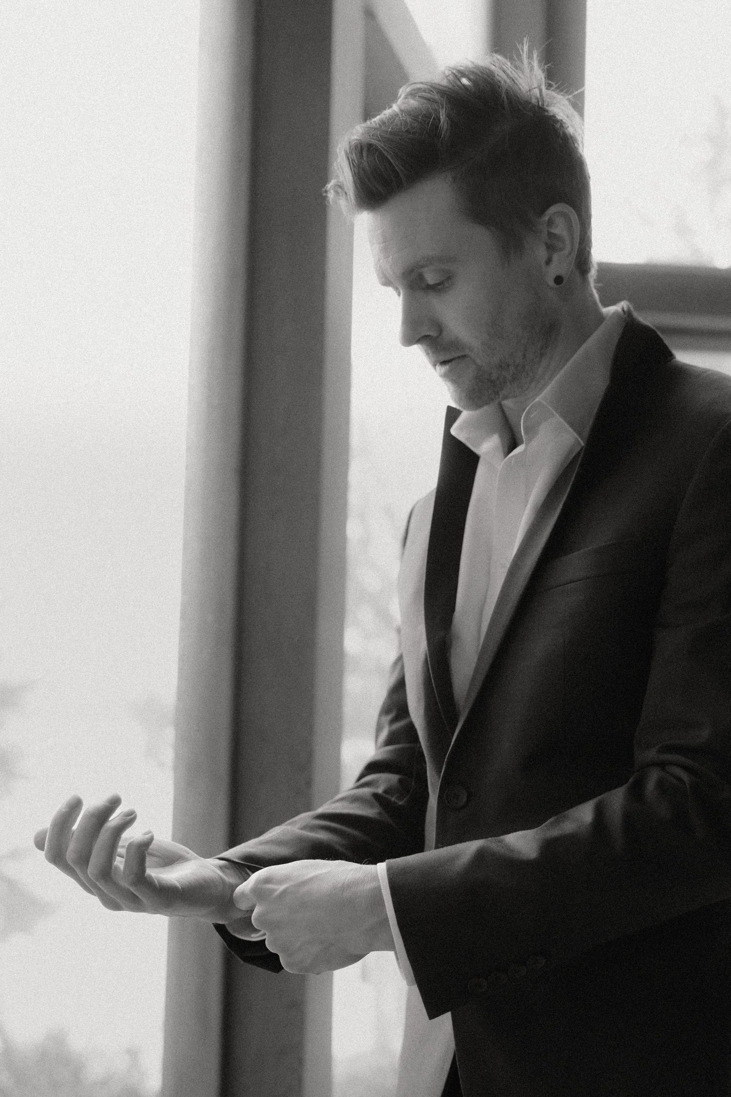 A person in a black suit adjusts their tie while standing near a window in black and white.