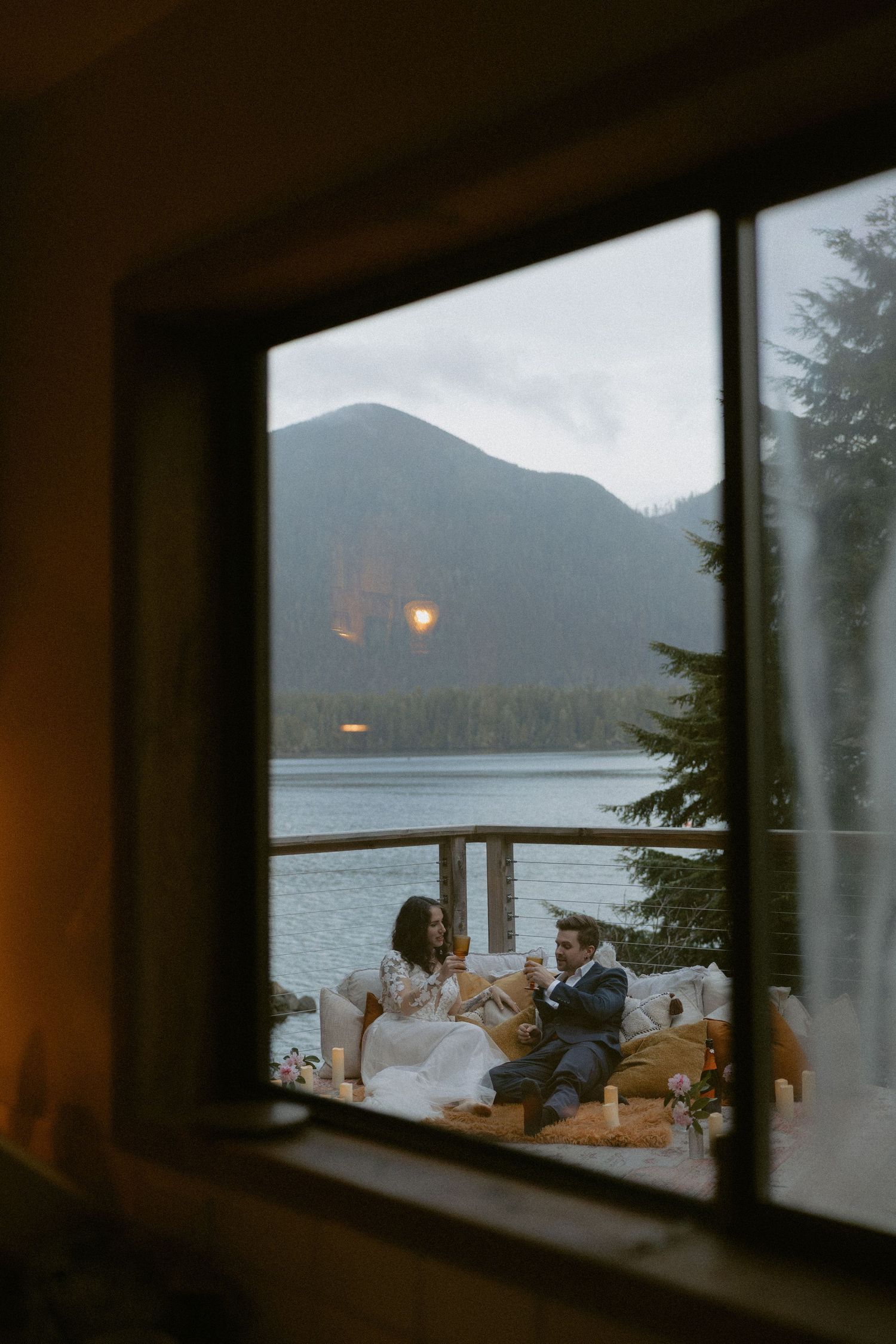 A cozy dinner scene viewed through a window frame with mountains and lake in the background.