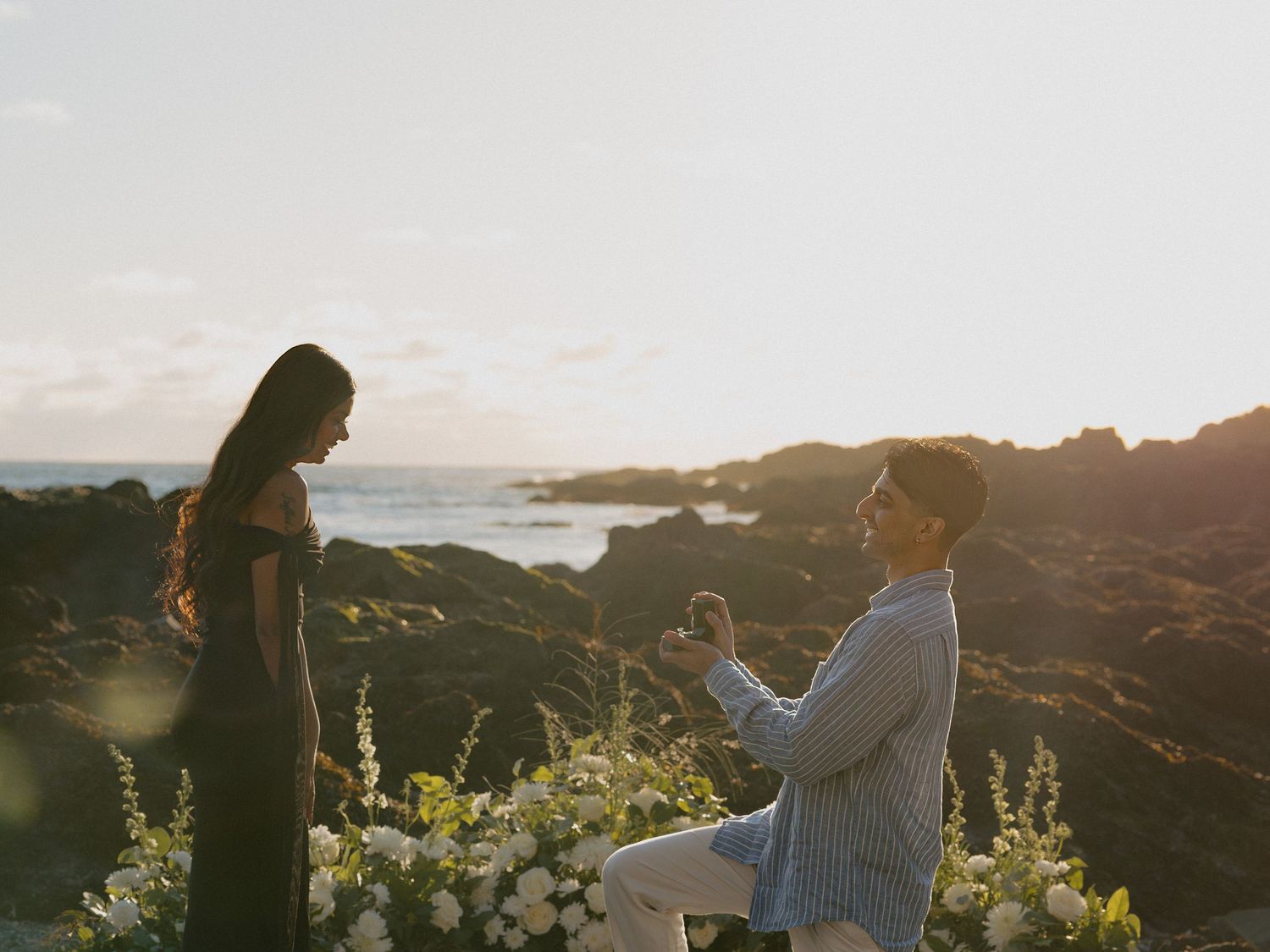Man kneeling to propose to woman at sunset on rocky coastal cliffside surrounded by white flowers.