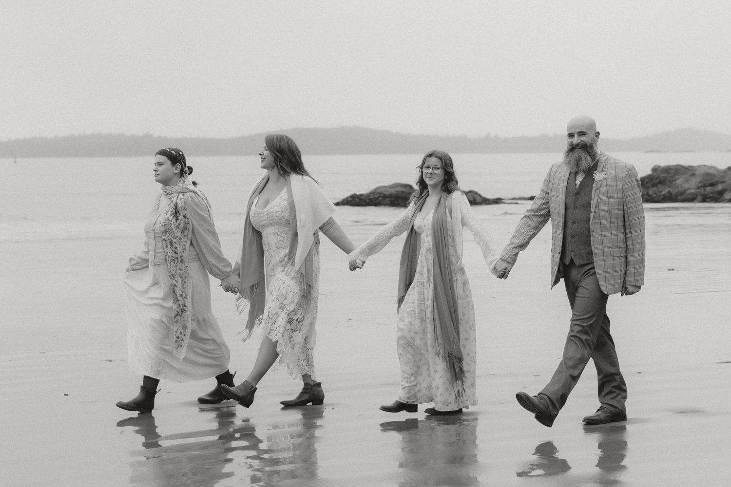 A group of people walk in a line while holding hands on a misty beach in black and white.