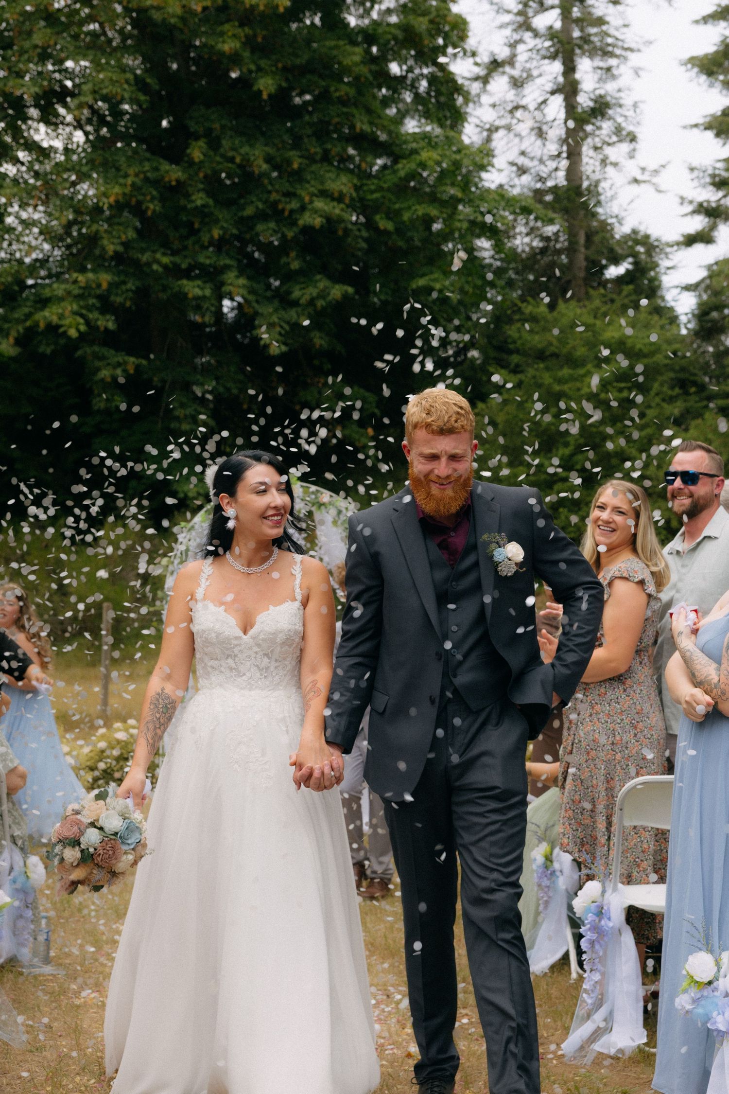 Bride and groom walk through confetti shower at outdoor wedding ceremony, surrounded by smiling guests.