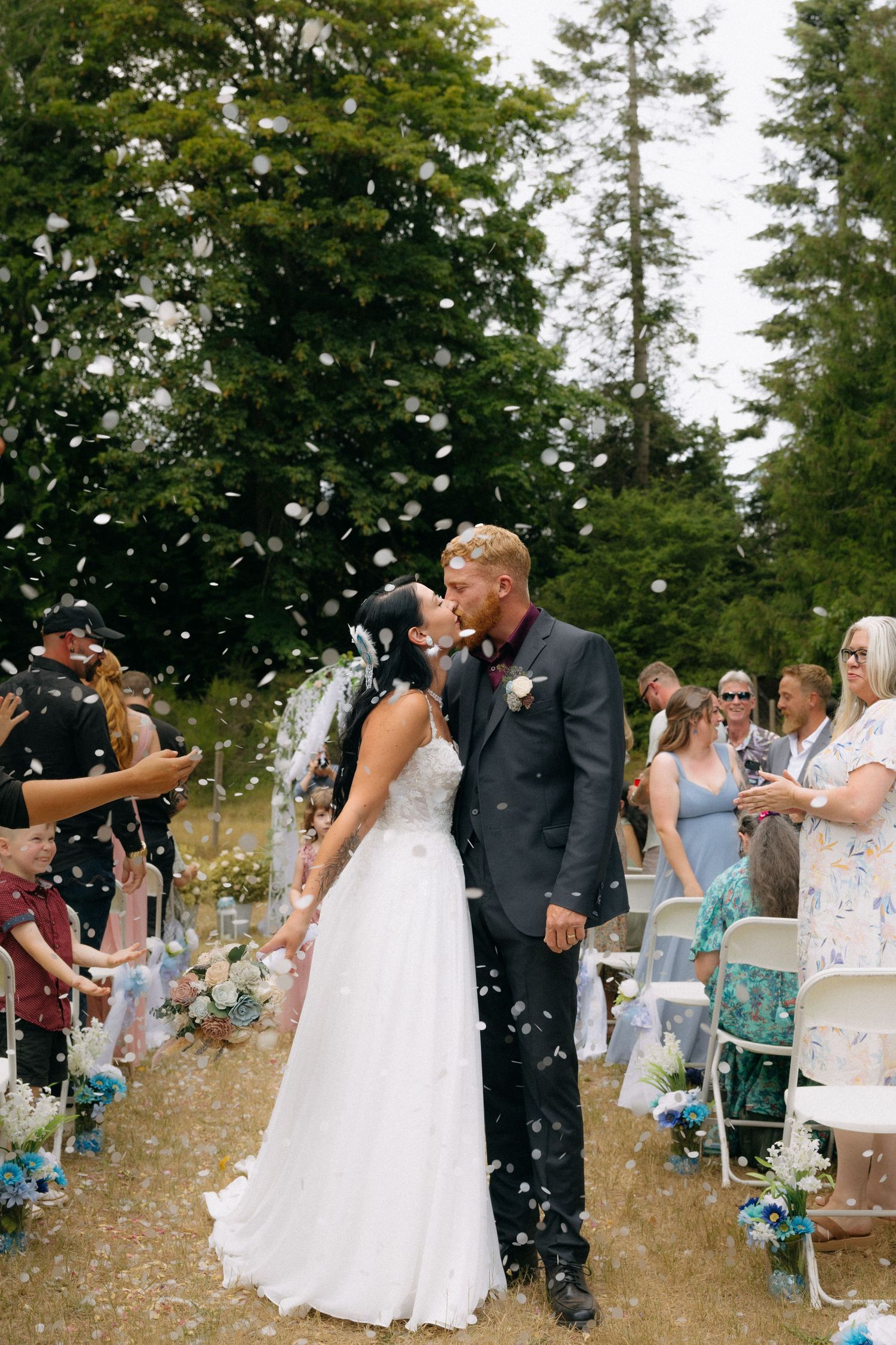 Bride and groom kissing during confetti exit at outdoor wedding ceremony surrounded by guests and tall trees.