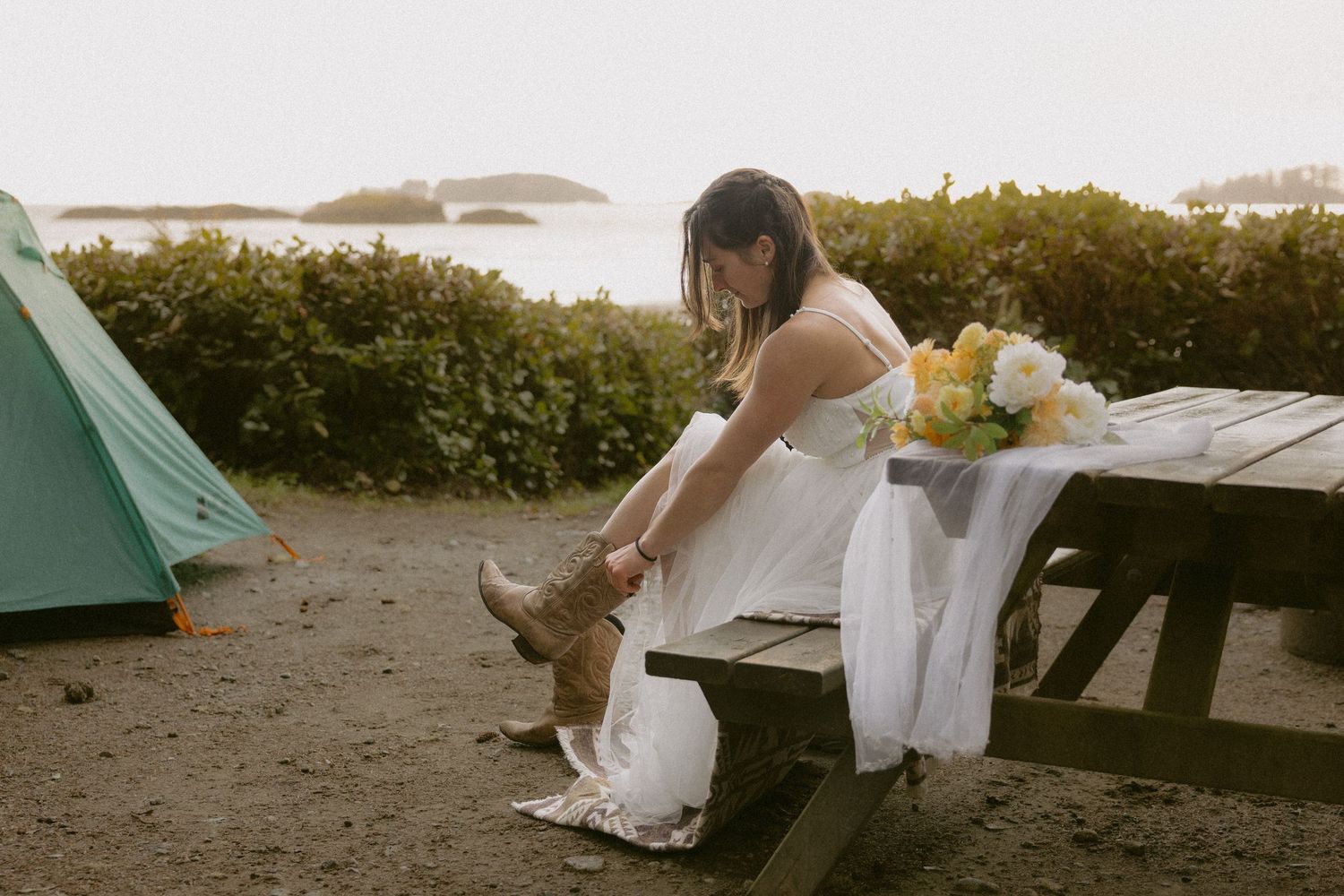 Tofino wedding bride putting on cowboy boots by her campsite at Bella Pacifica campground with her bouquet and veil on the picnic table