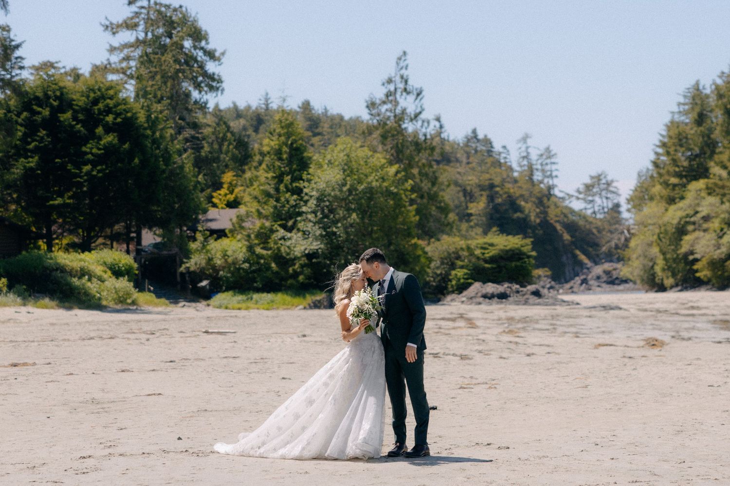Bride and groom share a romantic kiss on a sandy beach surrounded by lush green trees on a sunny wedding day.