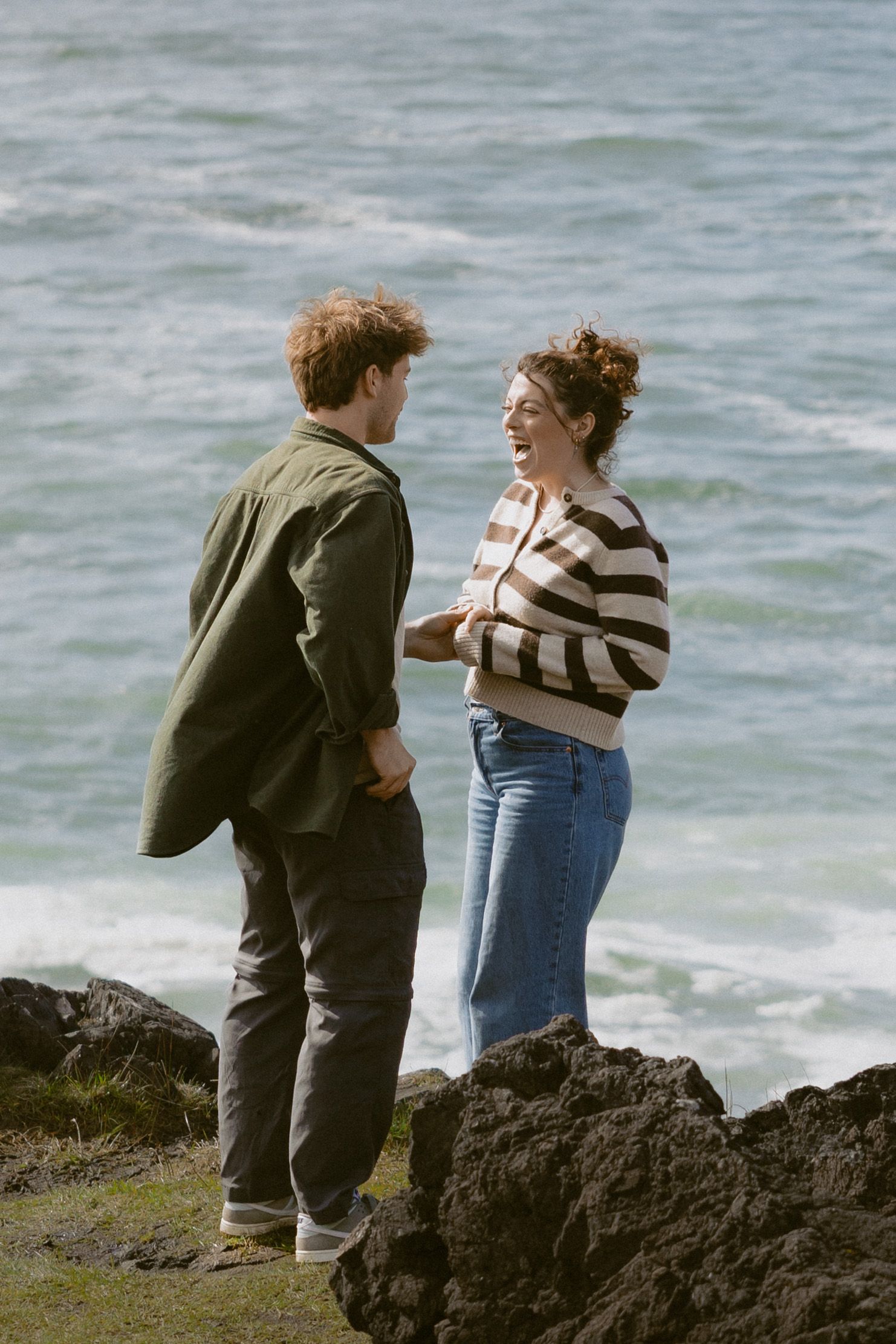Couple laughing together on rocky coastal cliffs with ocean waves in the background.