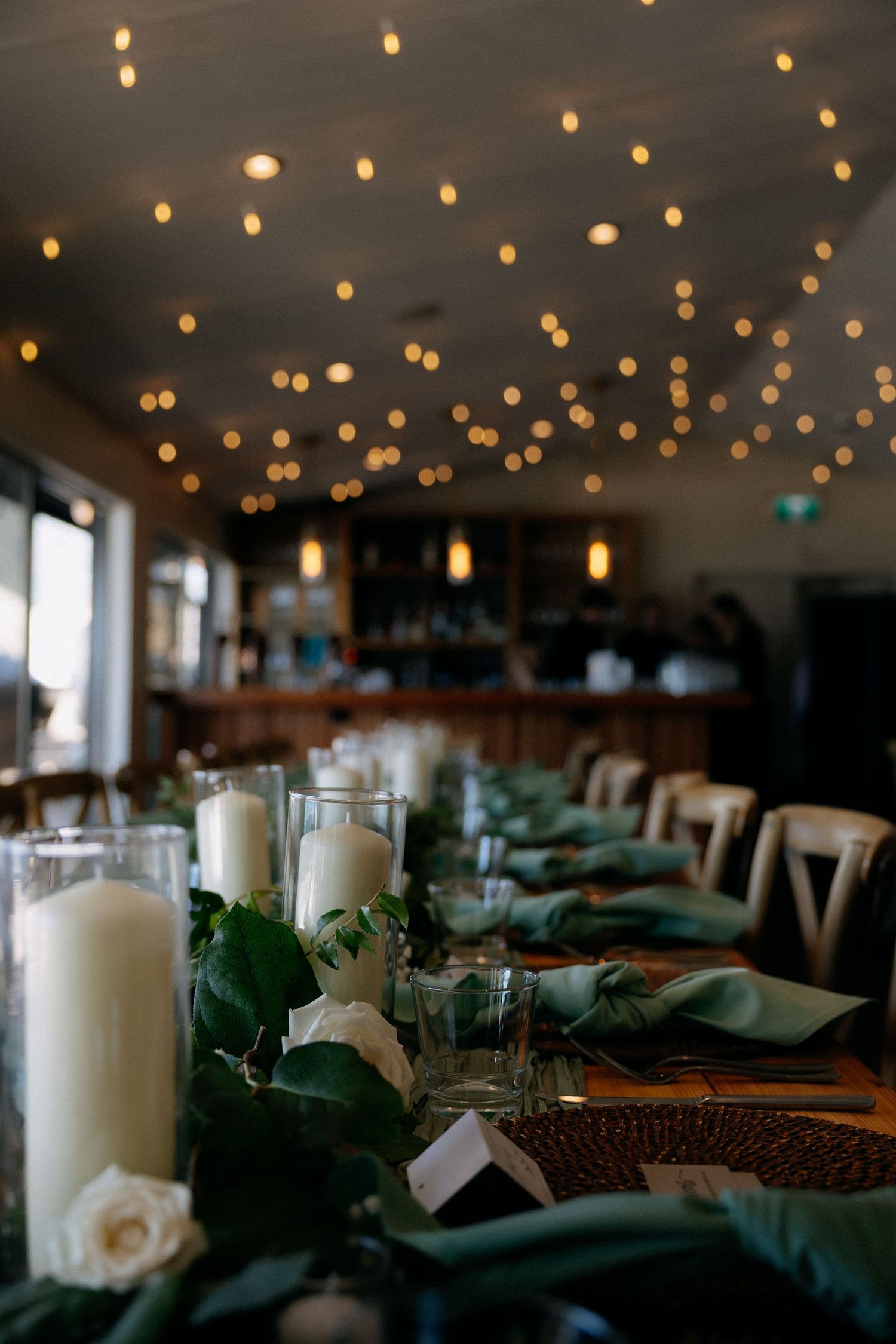 Elegant wedding reception table with white candles, green napkins, and bokeh string lights overhead.