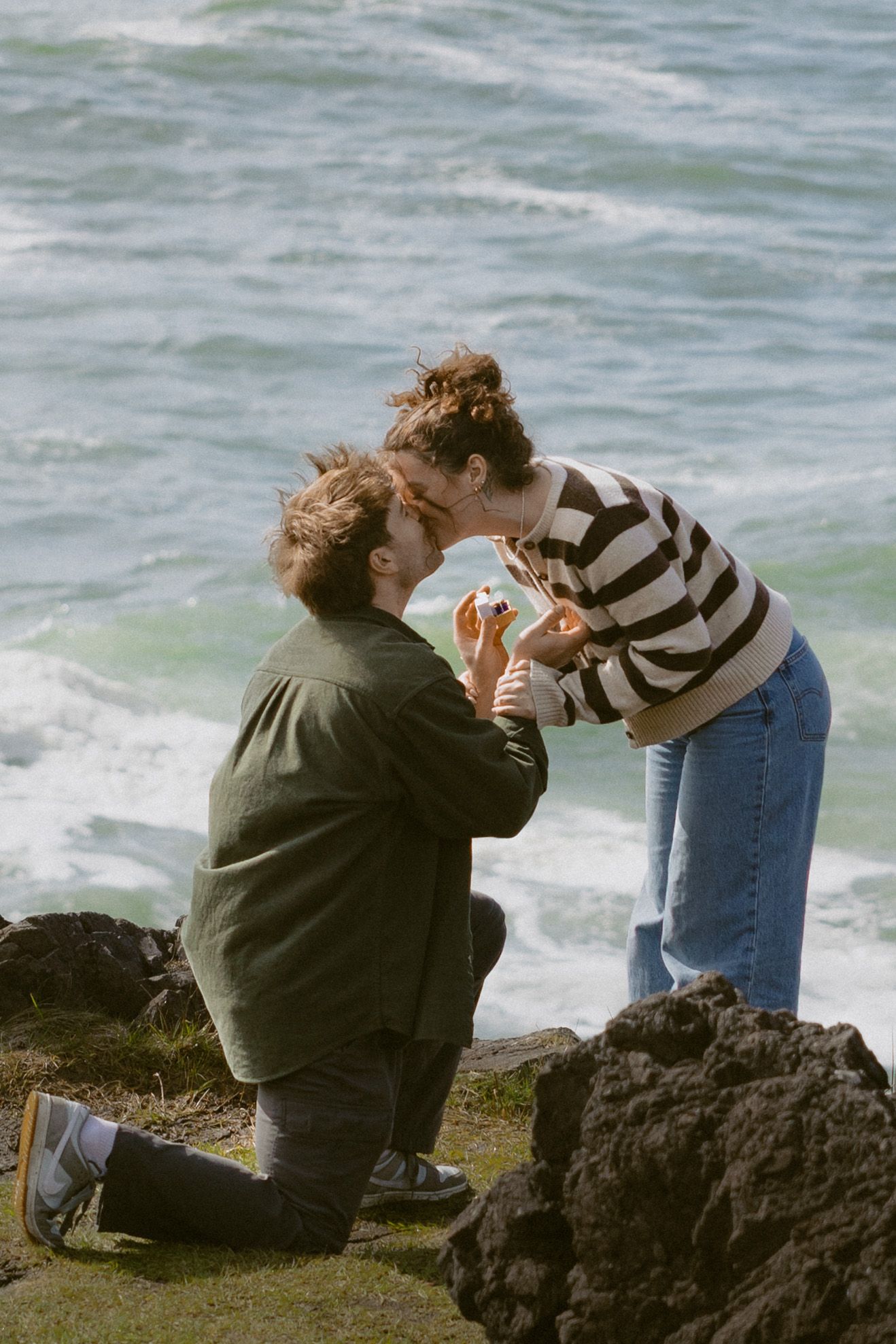 Man kneeling on rocky seaside cliff proposing to woman in striped shirt with ocean waves in background.