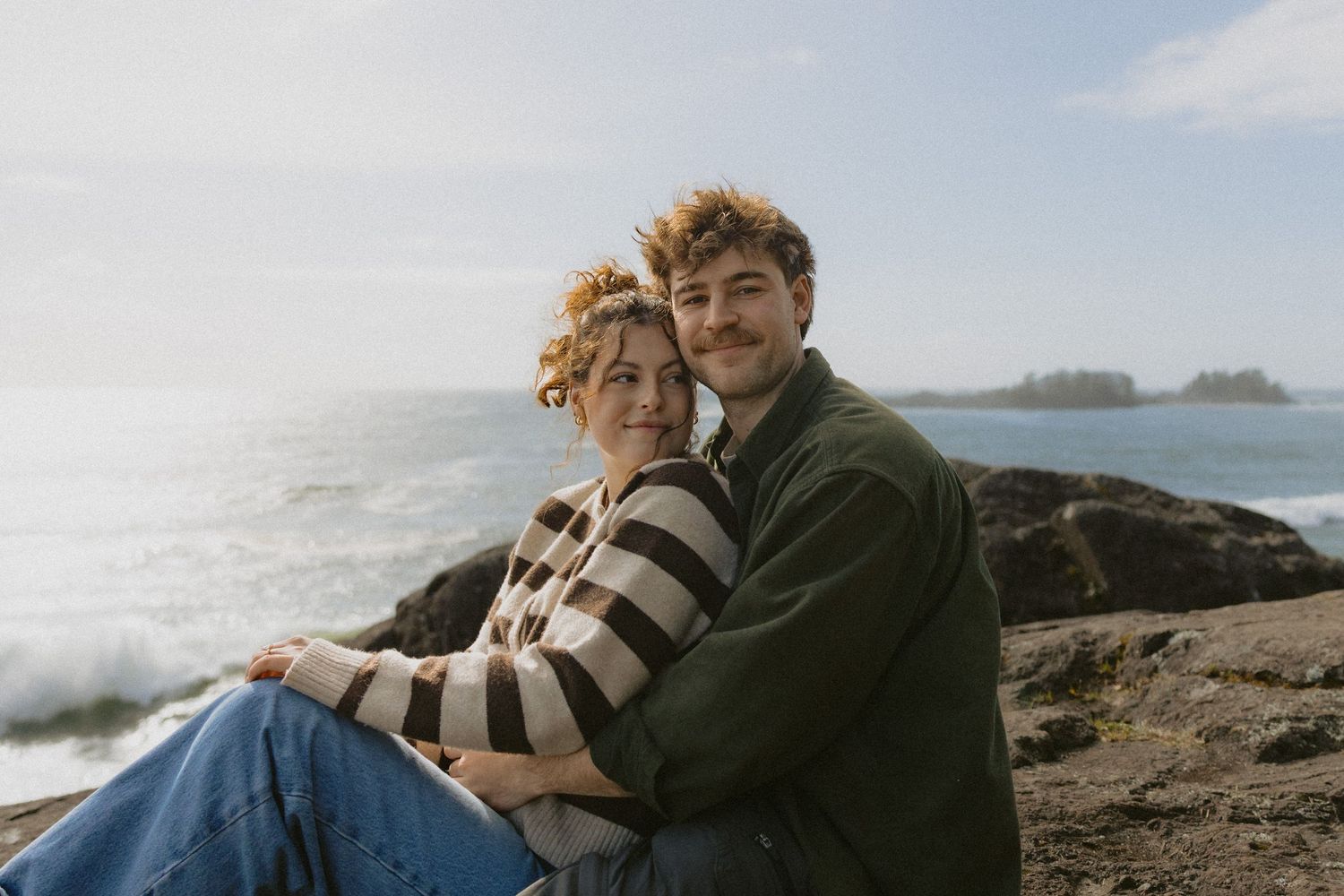 Couple sitting on rocky coastal shore embracing, woman in striped sweater leaning on man in green jacket.