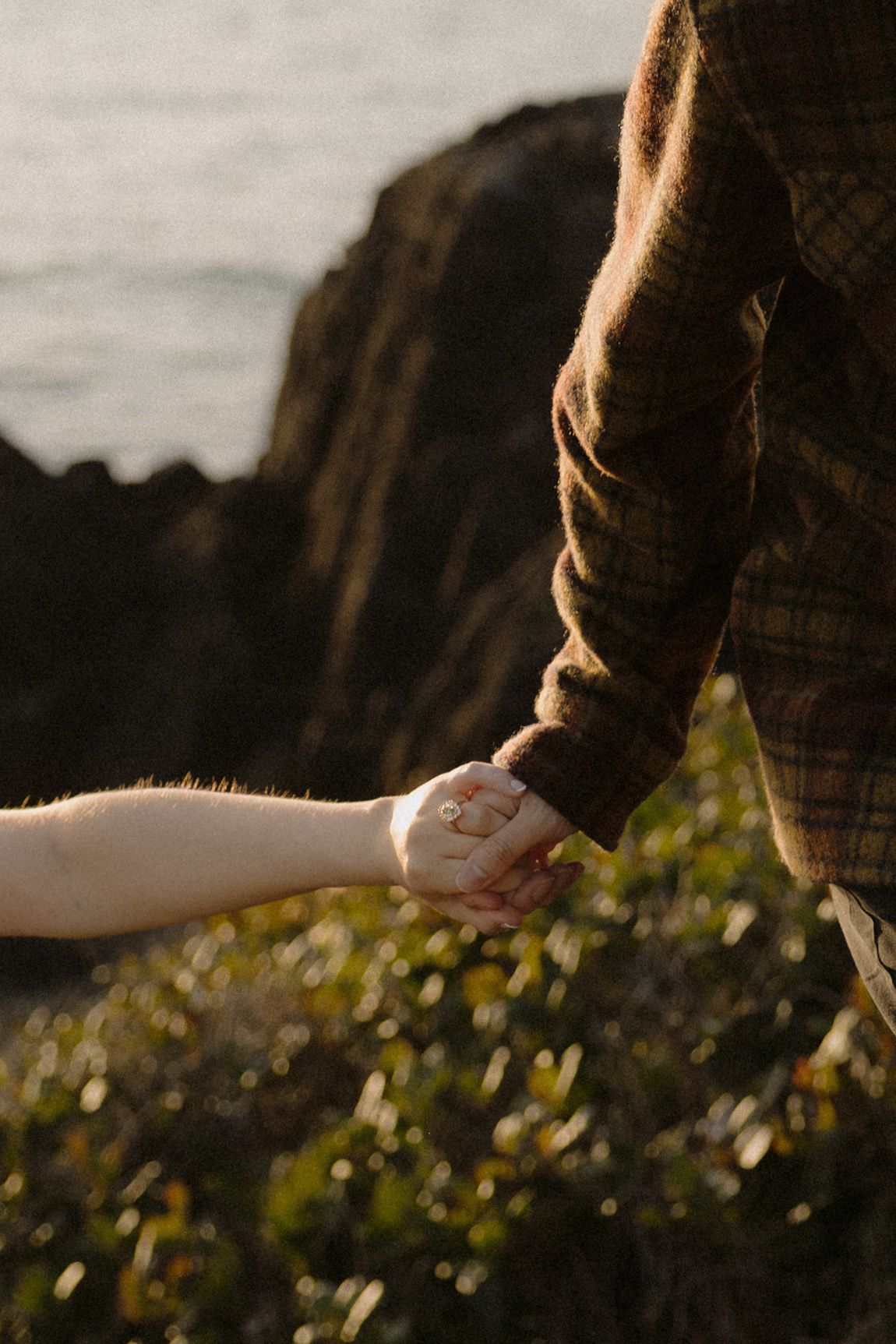 Close-up of two people holding hands near rocky coastal cliffs, one wearing a brown plaid jacket, with an engagement ring visible.